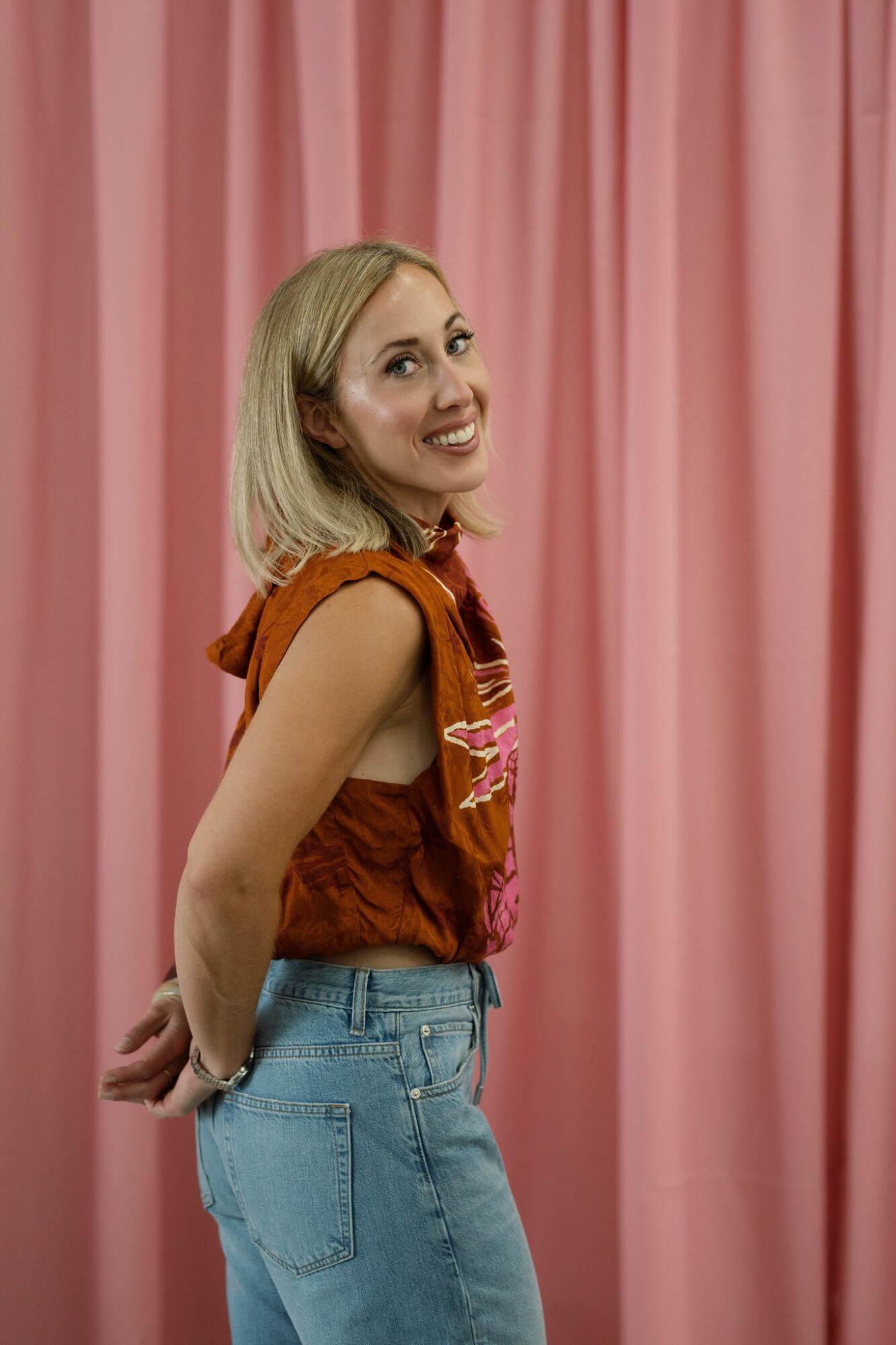 Young woman with blonde hair smiling, wearing a sleeveless orange top and blue jeans, standing in front of pink curtains.