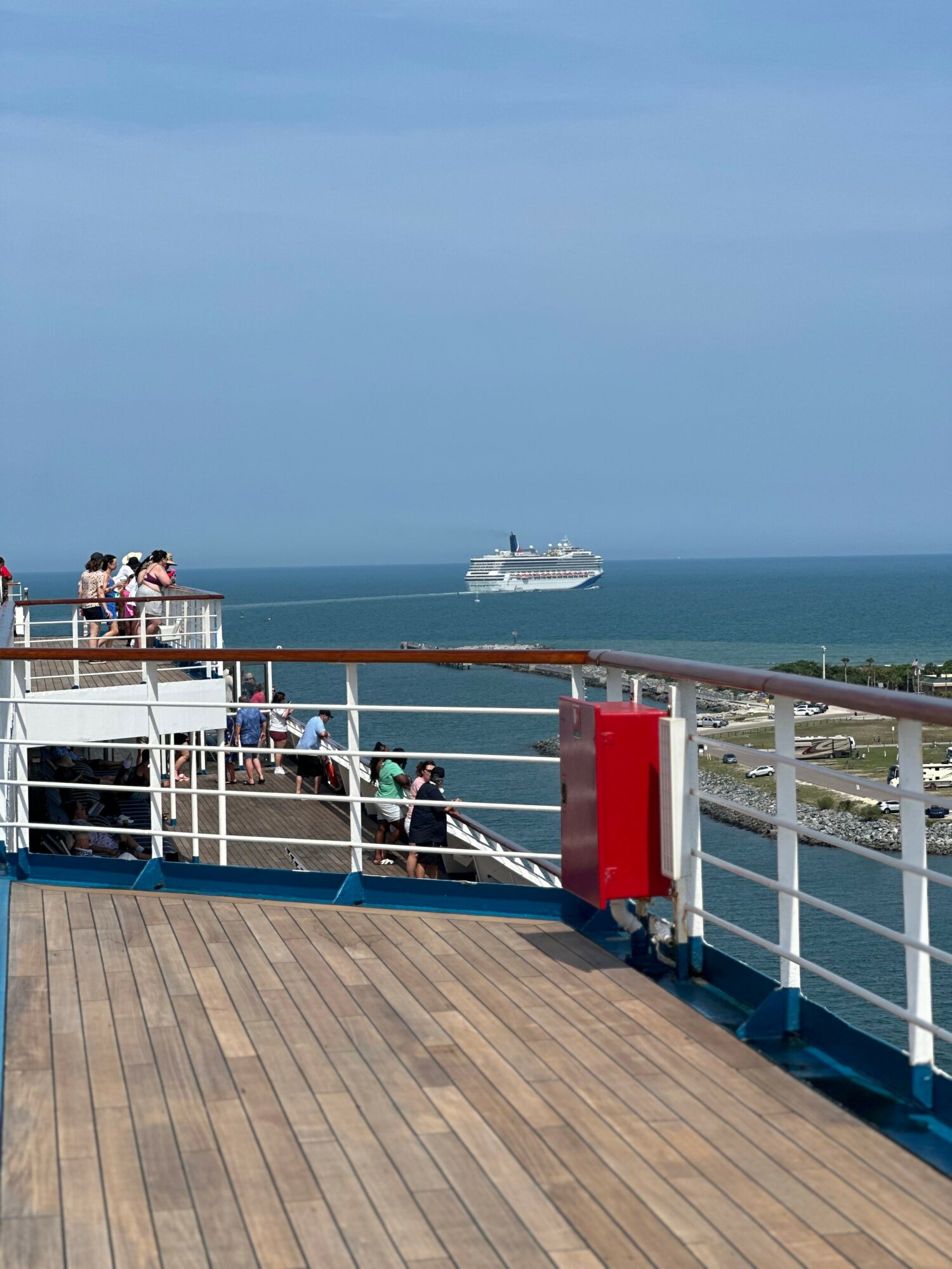 View from a ship's deck showing people, a railing, and a distant cruise ship on the ocean with a cloudy sky.