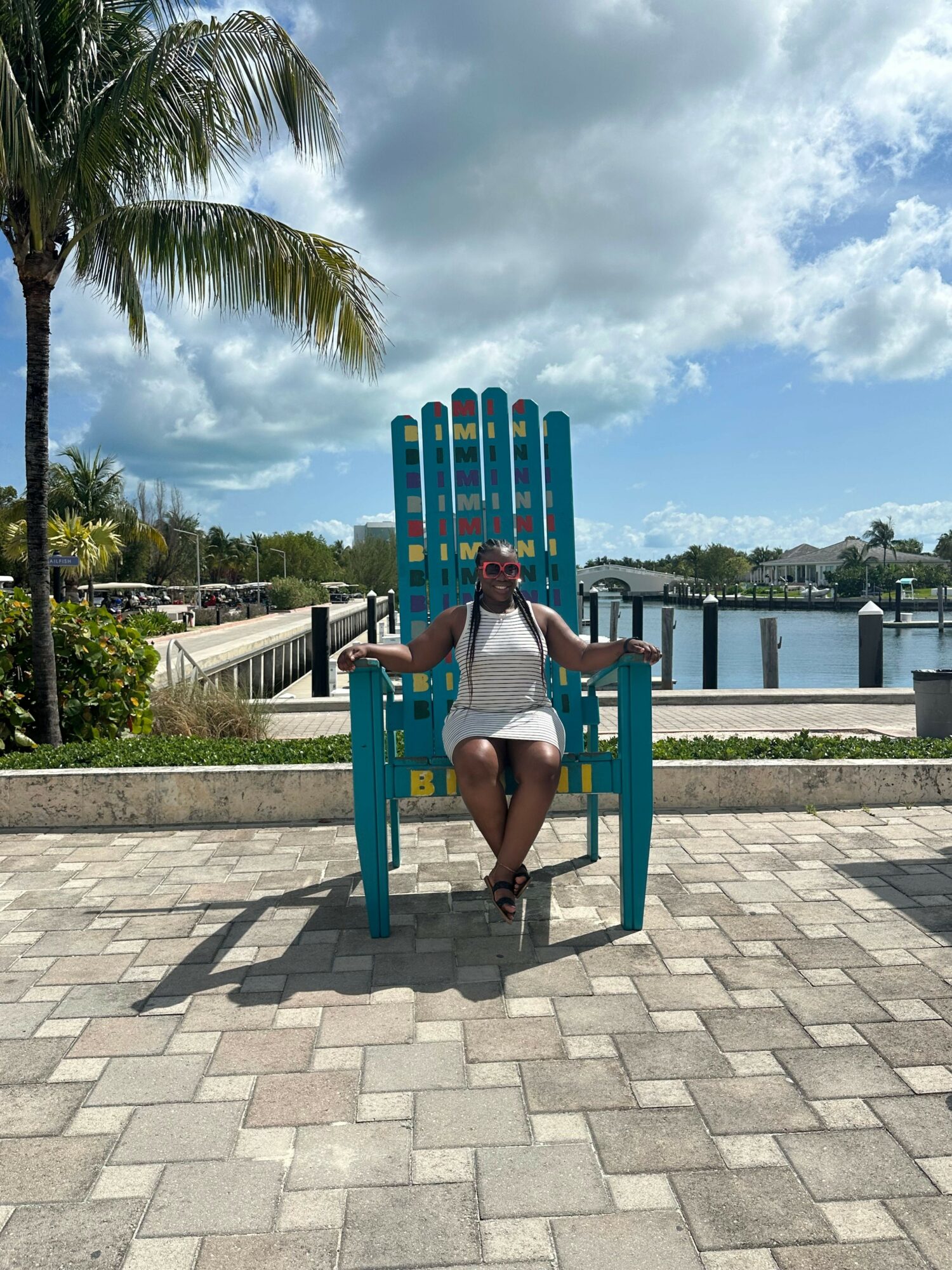 Person sitting on a large blue and green outdoor sculpture, with a palm tree and a bridge in the background, under a partly cloudy sky.