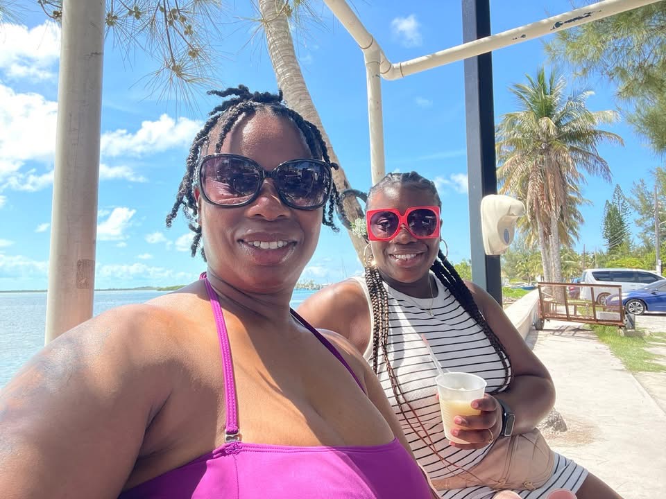 Two women smiling outdoors, one wearing sunglasses and a purple top, the other with red sunglasses and striped shirt, holding a drink.