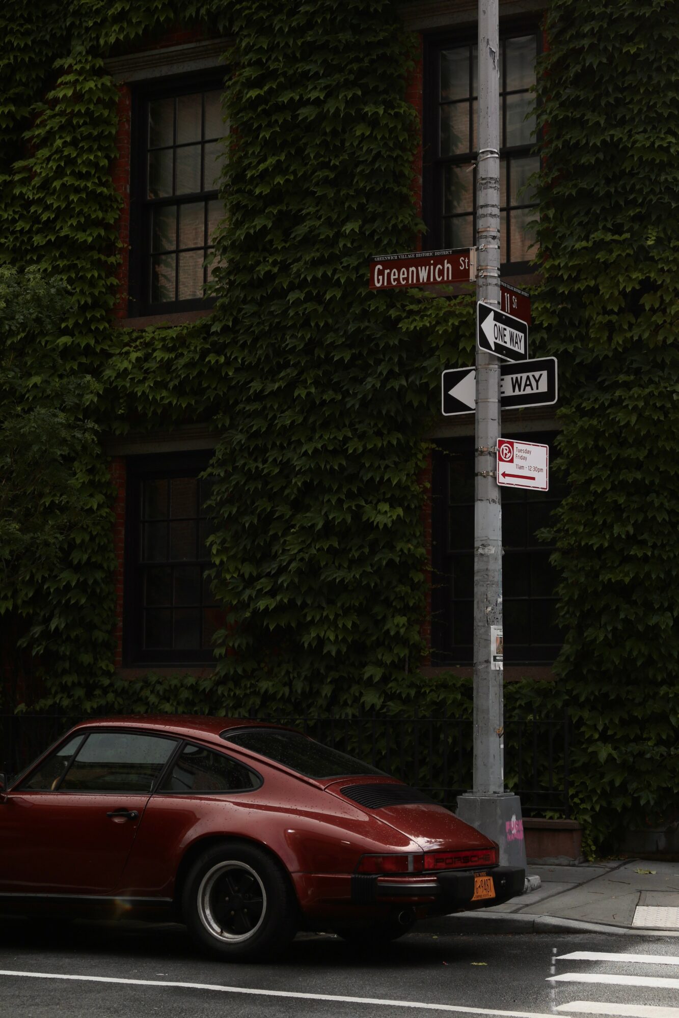 Red car parked on street next to a pole with street signs, in front of a building covered in green ivy.
