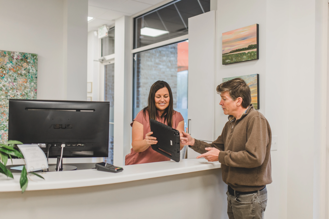 Two women at a reception desk, one holding a tablet, in a bright office with artwork on the wall.