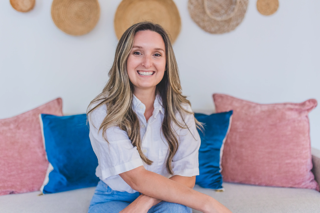 Smiling woman with long hair sitting on a sofa with pink and blue pillows, wall decor behind her.