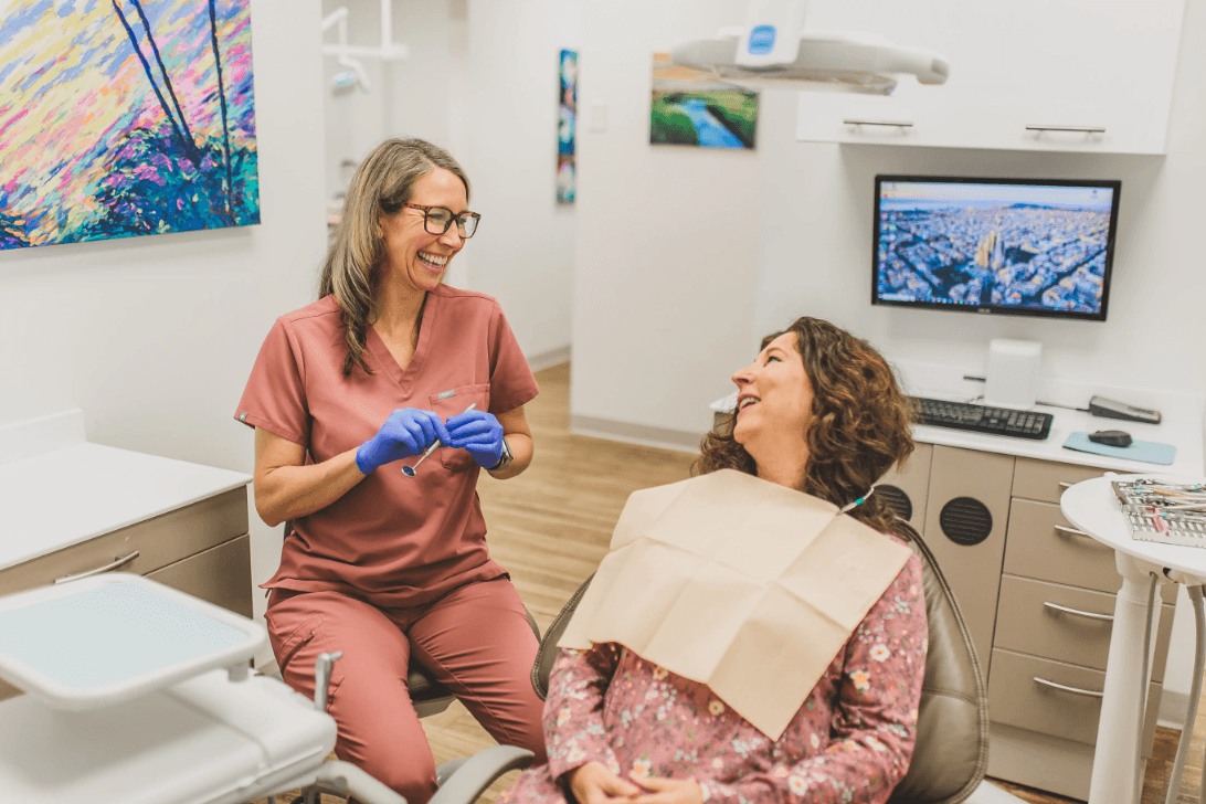 Dental professional smiling and talking to a patient in a dental office, patient sitting in chair with bib, professional wearing scrubs and gloves.