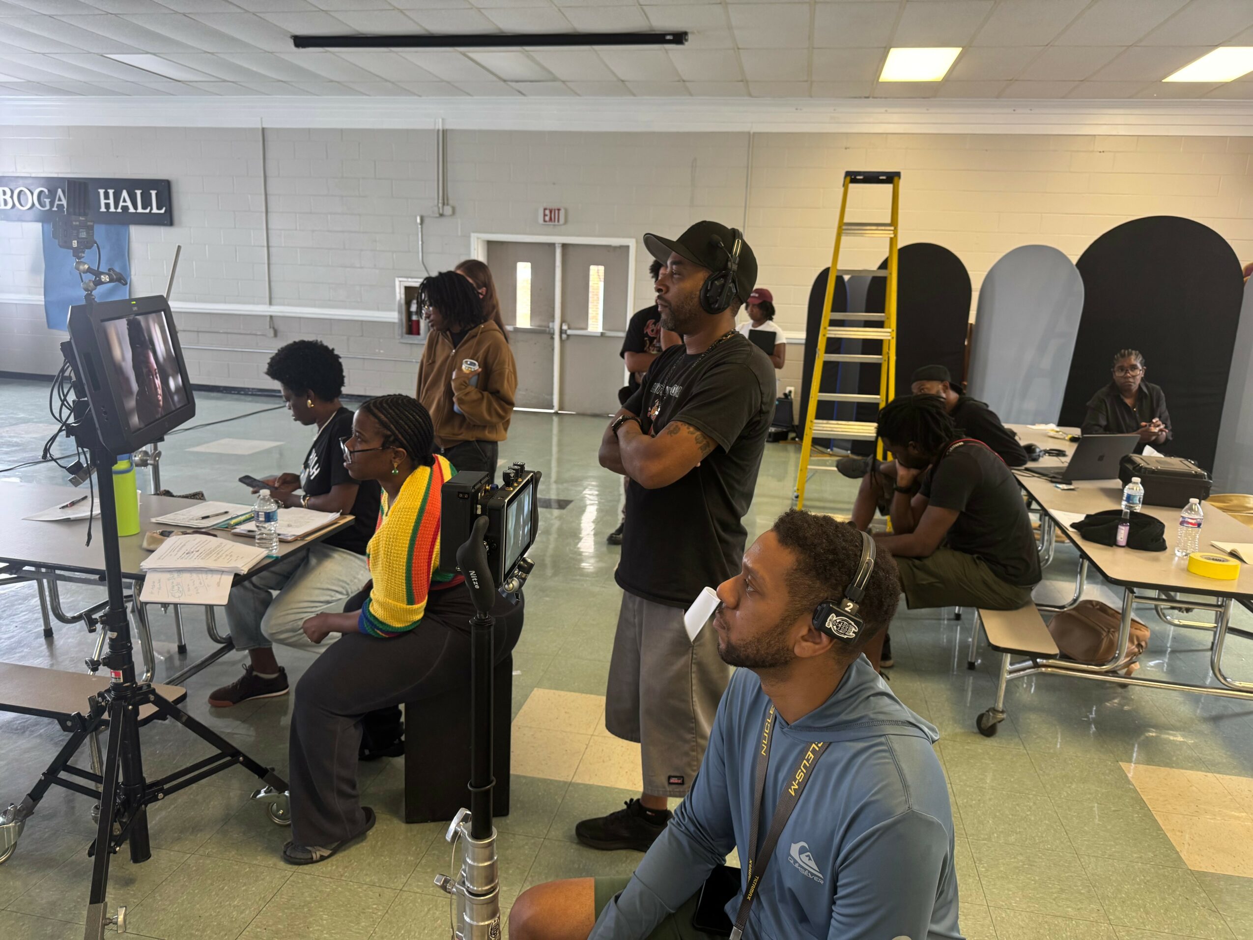 Group of people in a room with tables, chairs, and a ladder, some working on computers and others standing or sitting.