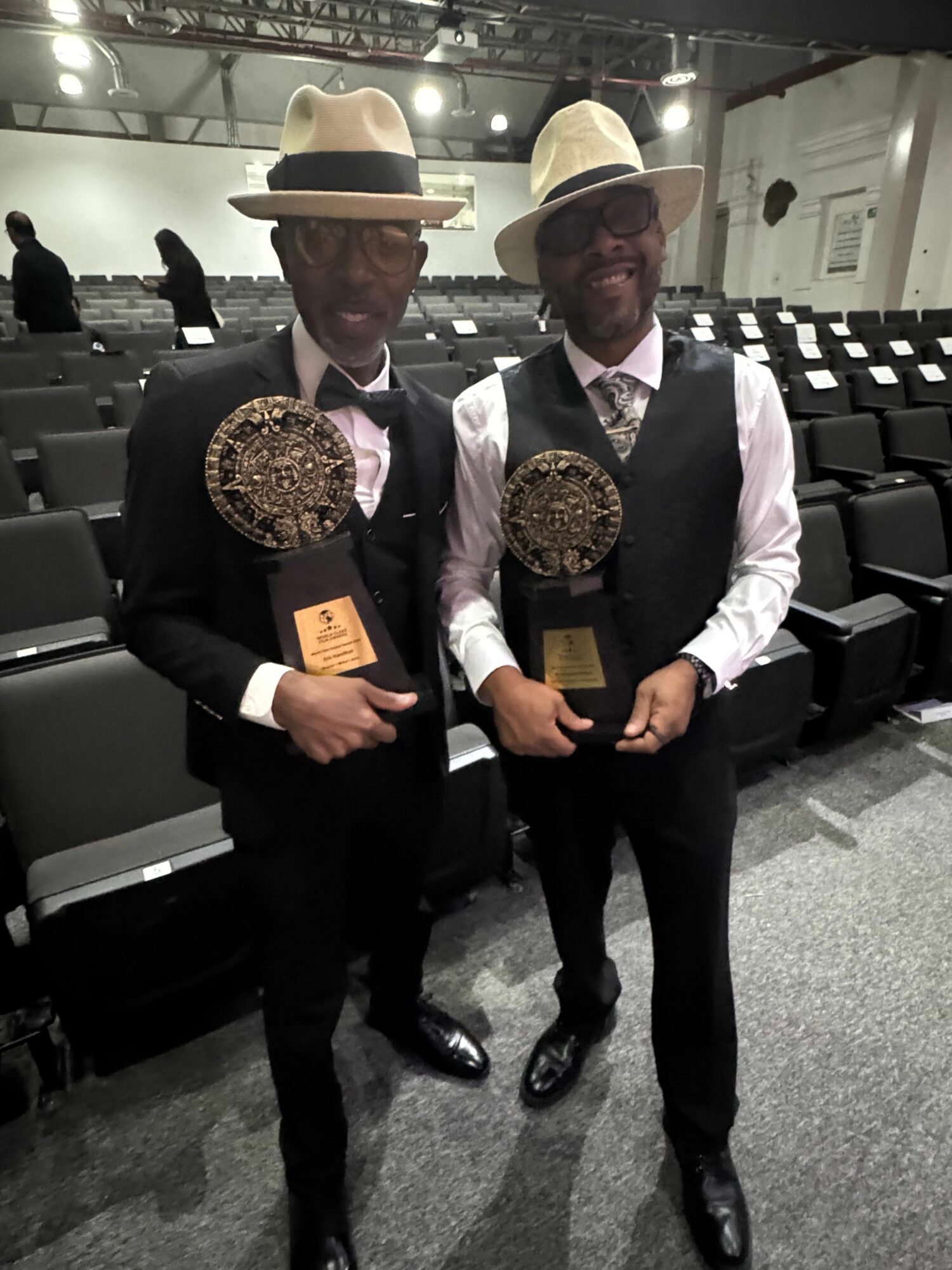 Two men in formal attire holding awards and certificates, standing in an indoor event space with black chairs.
