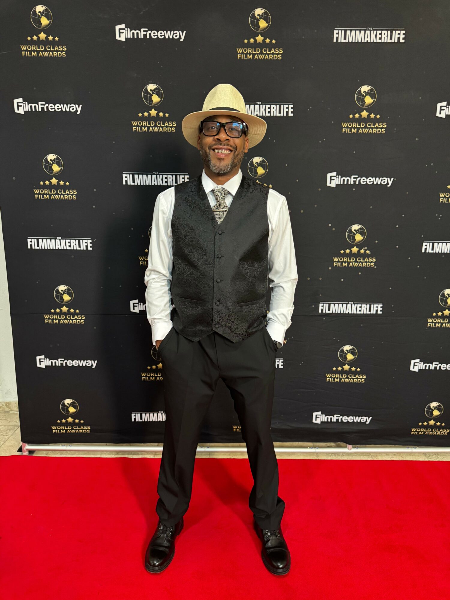 Man in a suit and hat standing on red carpet with black backdrop featuring gold logos and text, smiling at camera.