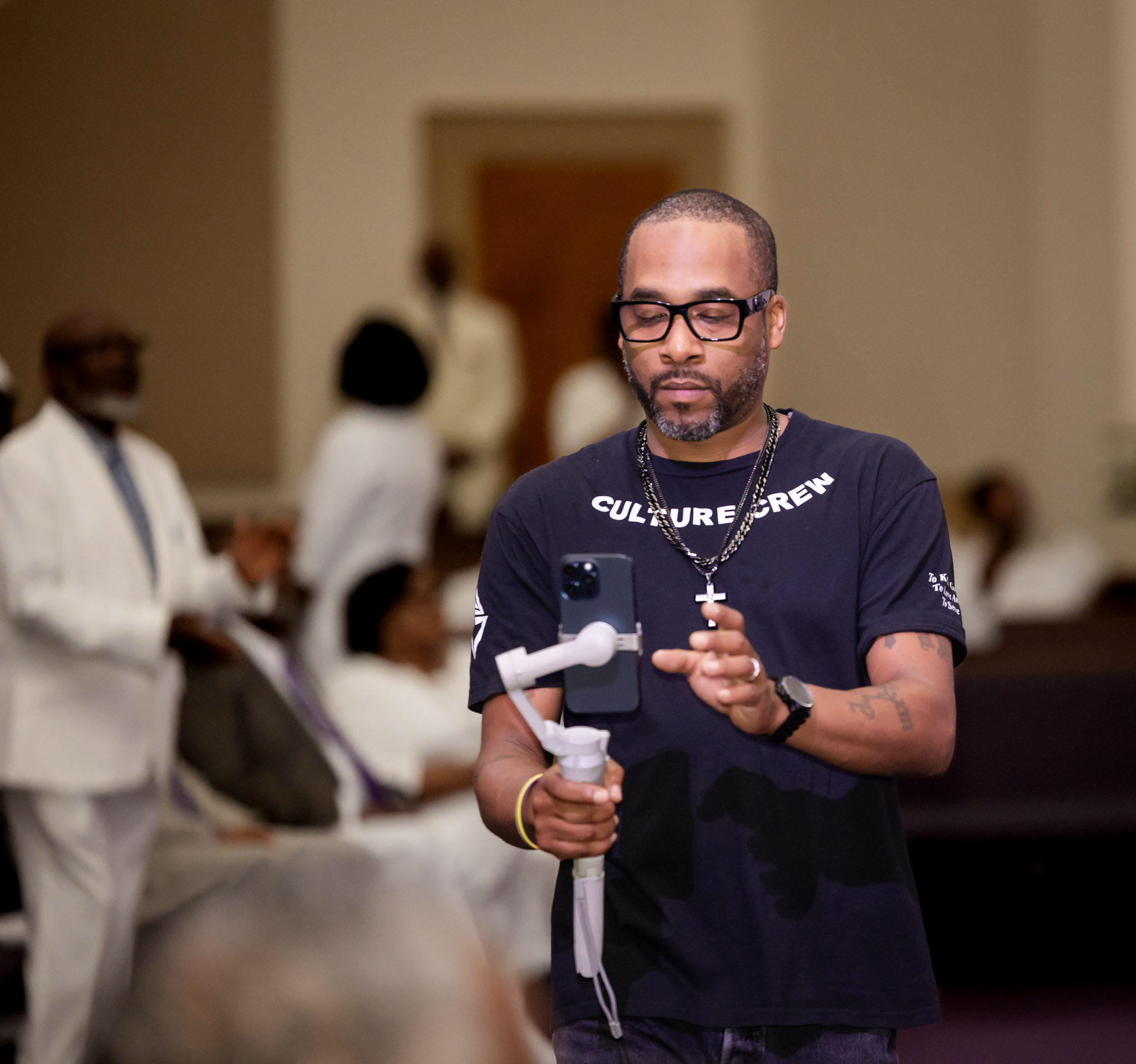 Man with glasses and a beard holding a camera, wearing a black T-shirt, in a room with people in the background.
