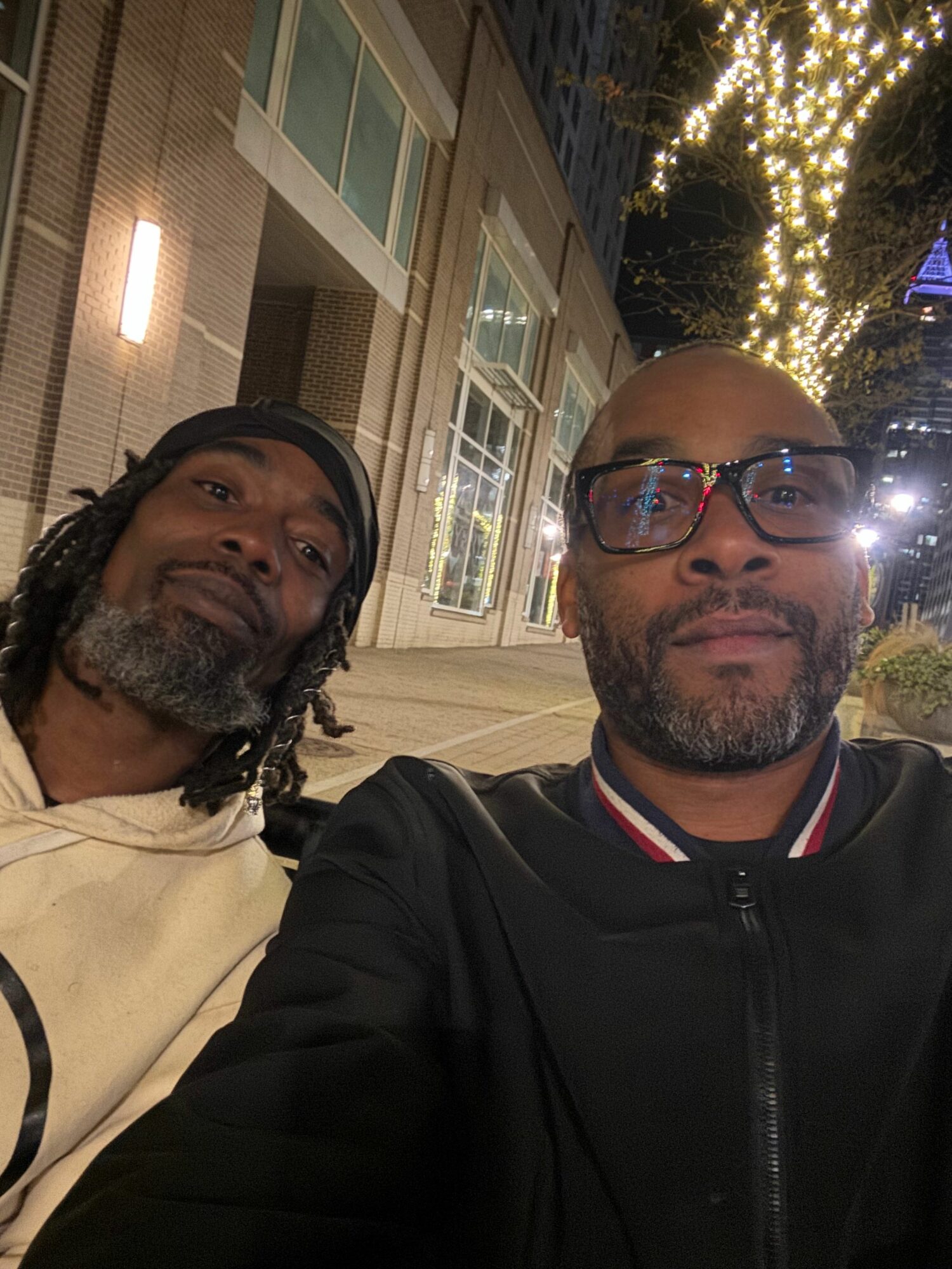 Two men with beards and glasses pose outdoors at night, illuminated by string lights, in front of a building with large windows.