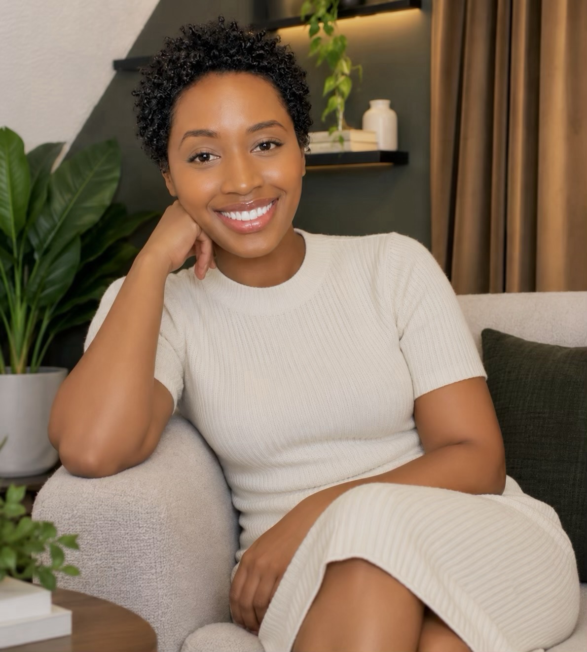 Smiling woman with short curly hair sitting on a sofa in a cozy room, wearing a cream-colored dress, with plants and curtains in background.