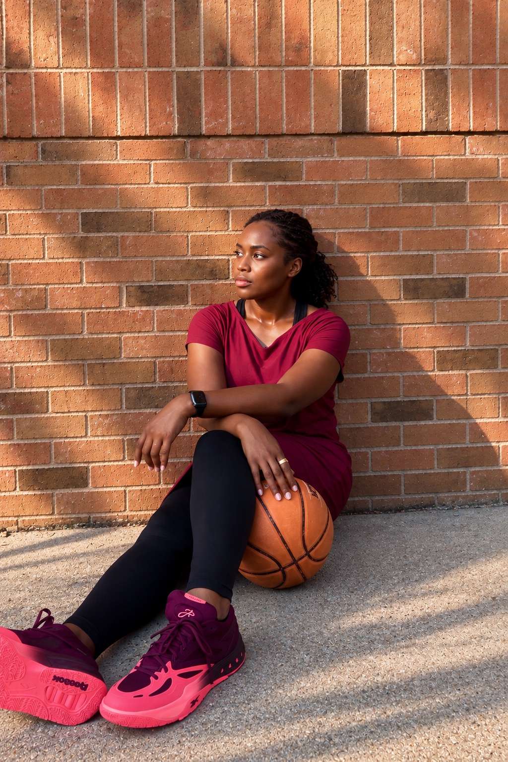 Young woman in a maroon shirt and black leggings sitting on a basketball against a brick wall, with shadows on the wall.