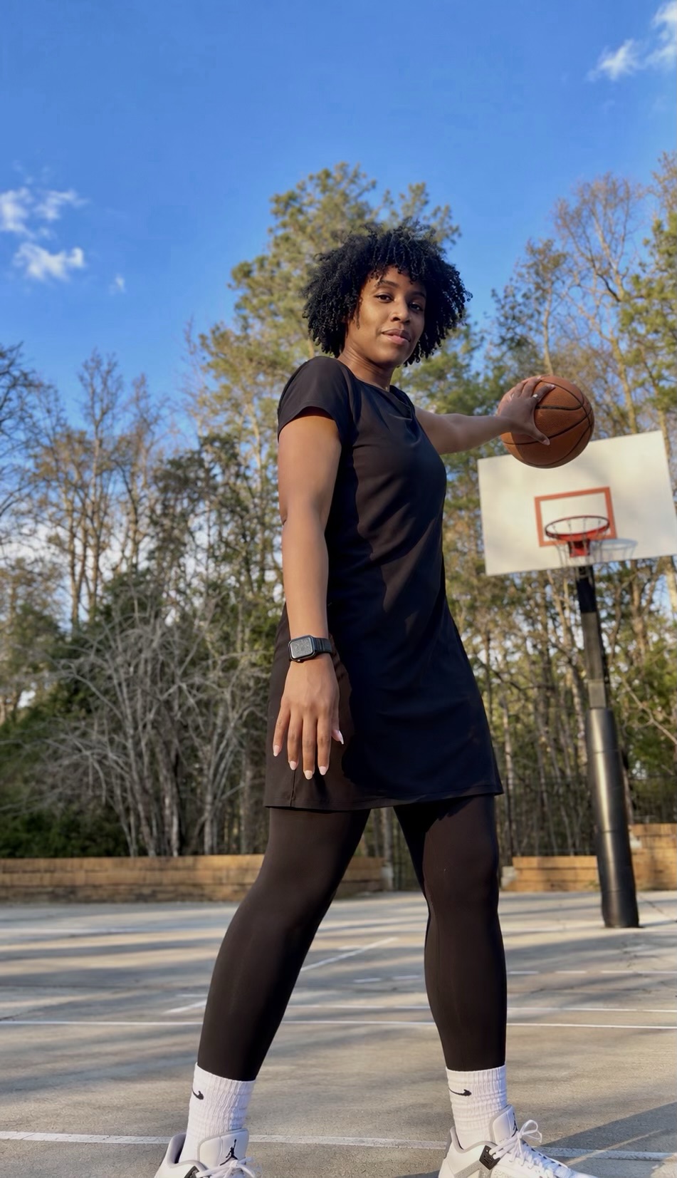 Woman standing outdoors on a basketball court holding a basketball, trees and blue sky in background.