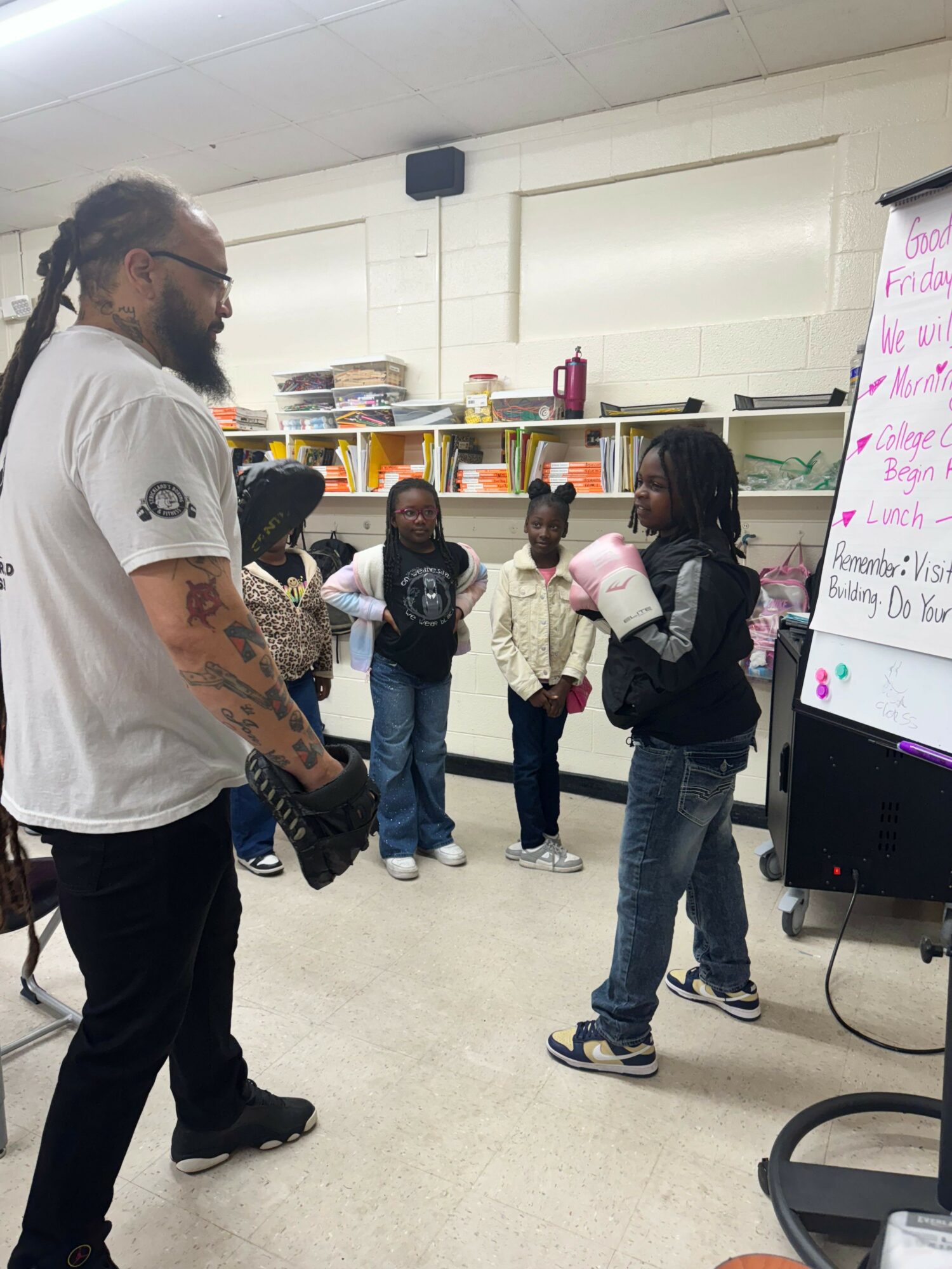 Group of people standing in a room with shelves and a whiteboard, some holding cups, a person with tattoos in the foreground.