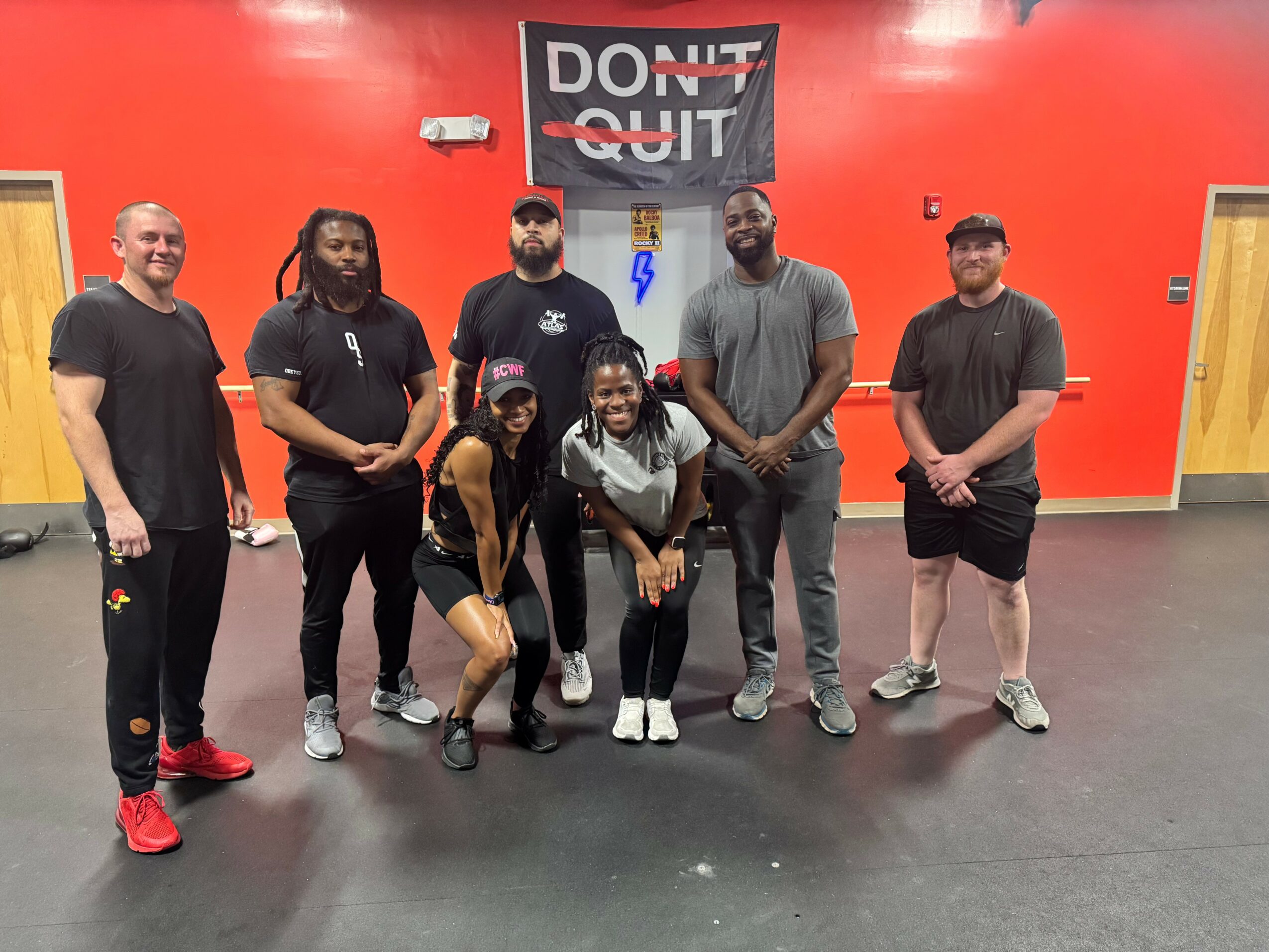Group of seven people standing in a gym with a red wall and a sign that says 'DON'T QUIT'.