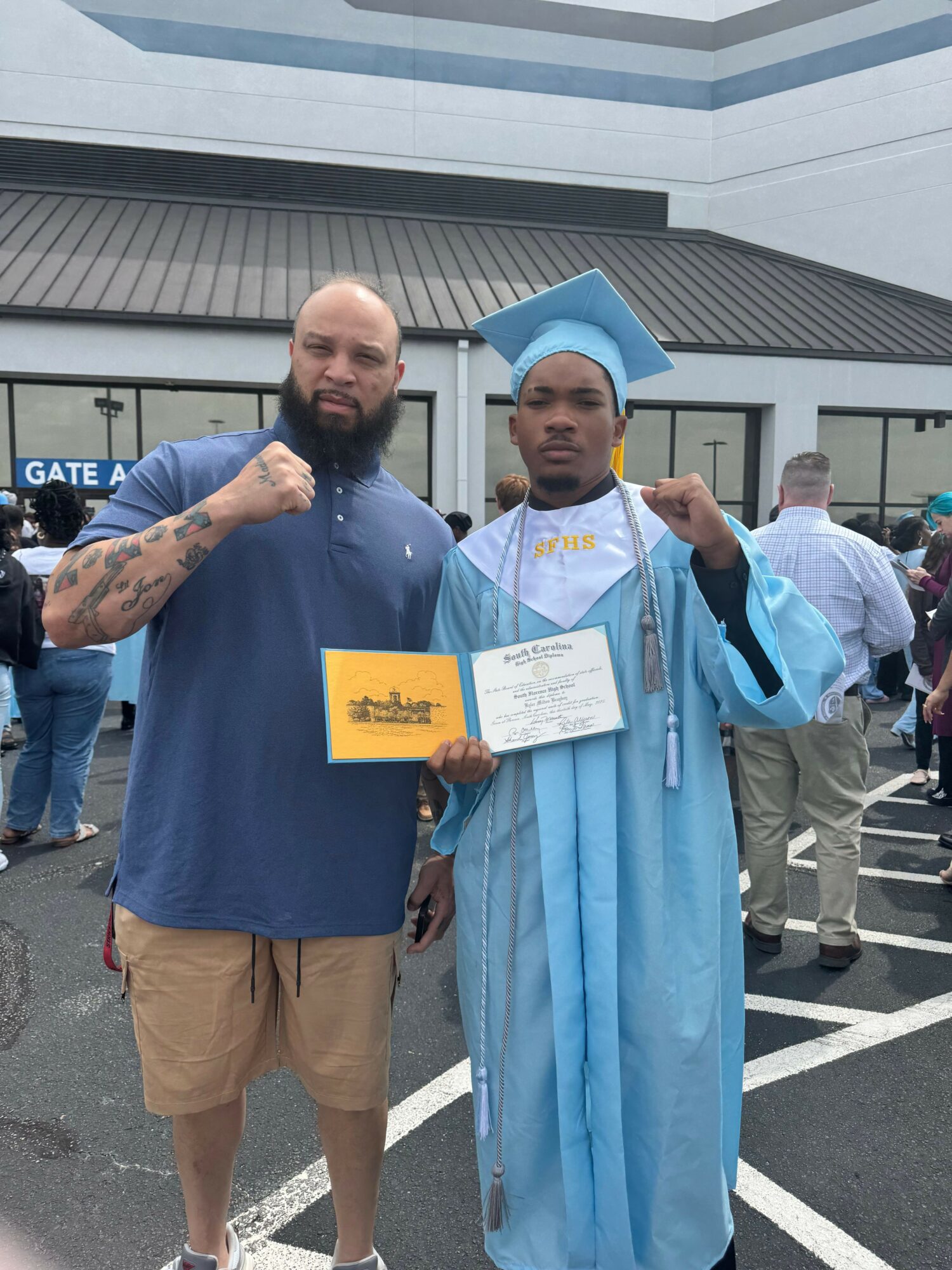 Graduate in cap and gown holding diploma, standing with a man in casual clothing outside a building.