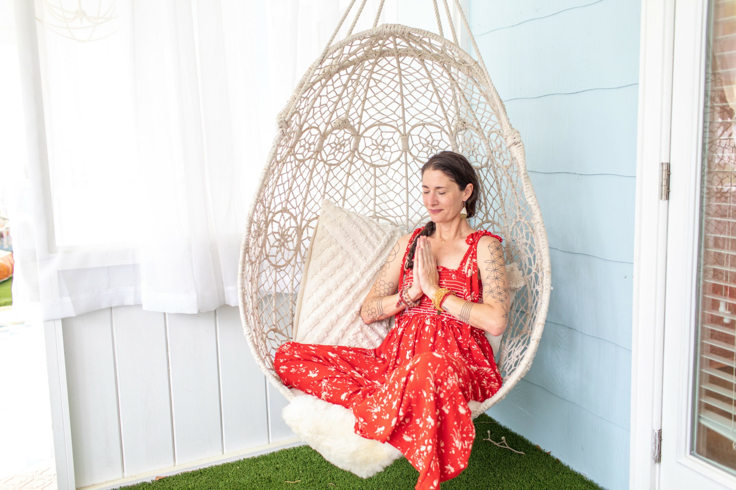 Woman in red dress sitting cross-legged in hanging chair with hands in prayer position, in bright room with white curtains and blue wall.