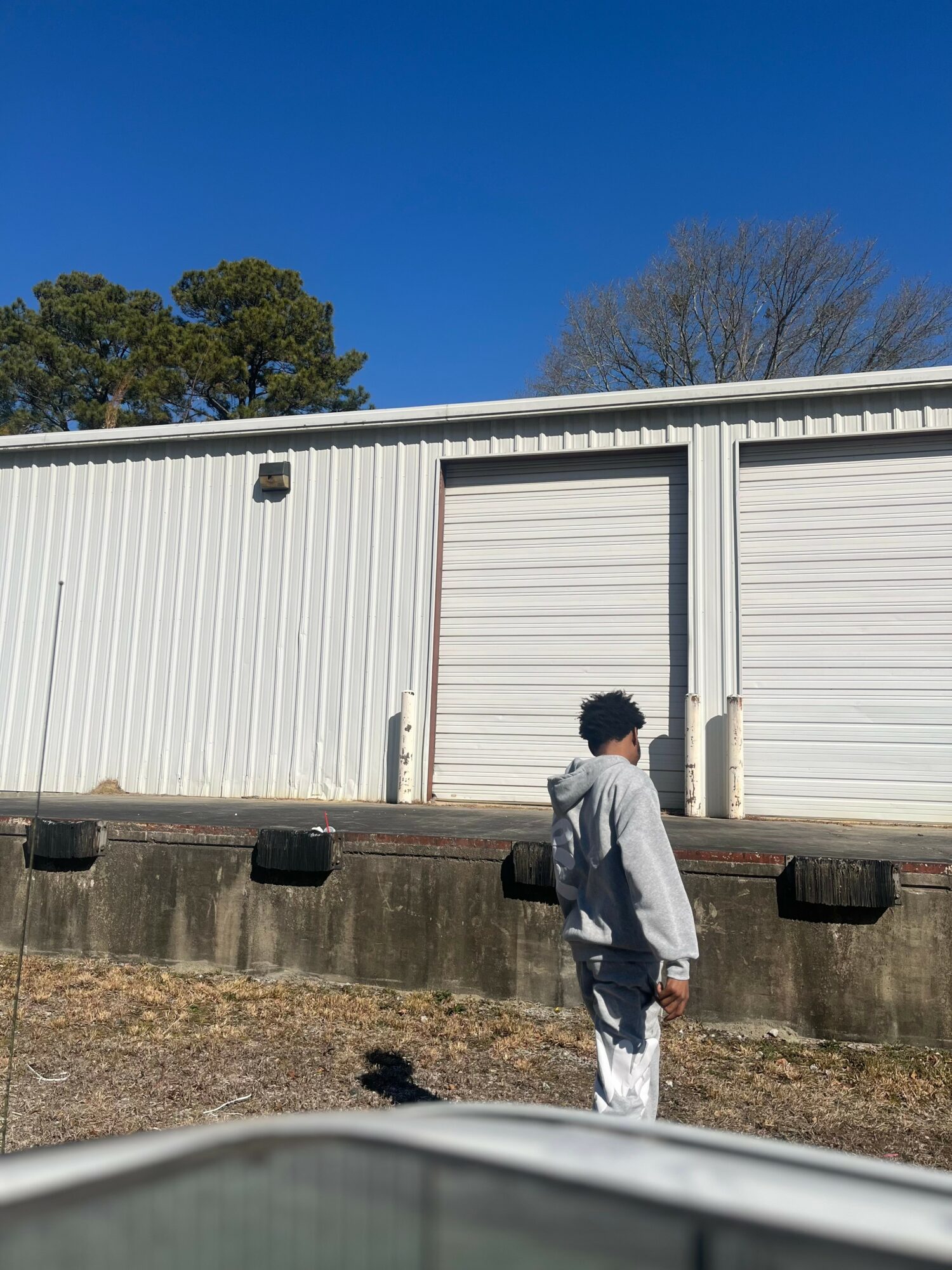 Person with curly hair wearing a light gray hoodie and white pants standing outside near a white industrial building with closed garage doors, under a clear blue sky.