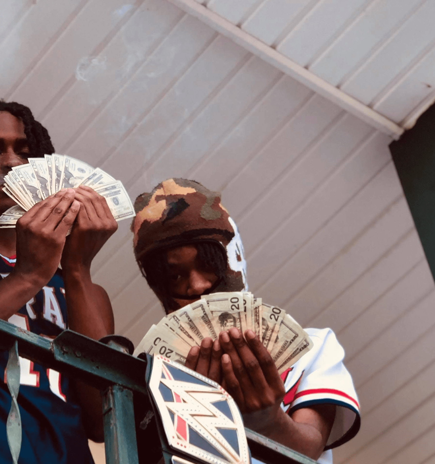 Two young men holding and displaying large amounts of cash, with a white ceiling in the background.