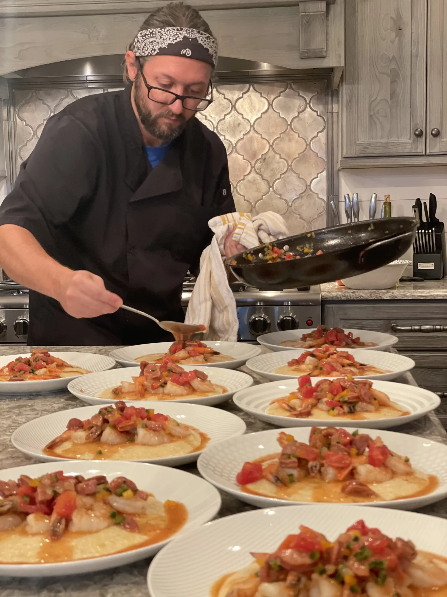 Chef in black uniform and headband cooking in kitchen, preparing multiple plates of food with toppings, on a countertop.