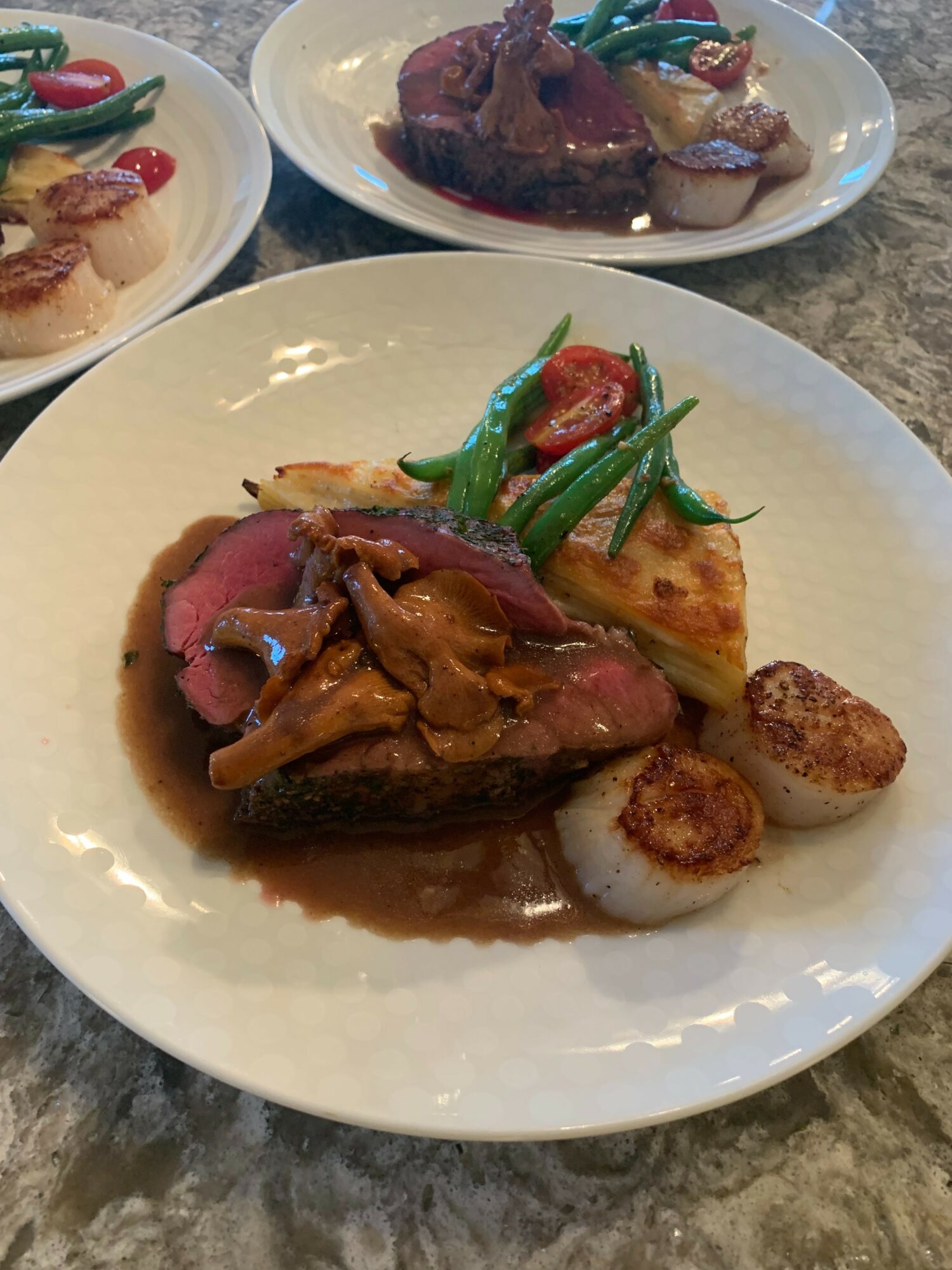 Plate with cooked meat, green beans, cherry tomatoes, potato slices, and seared scallops on a white dish.