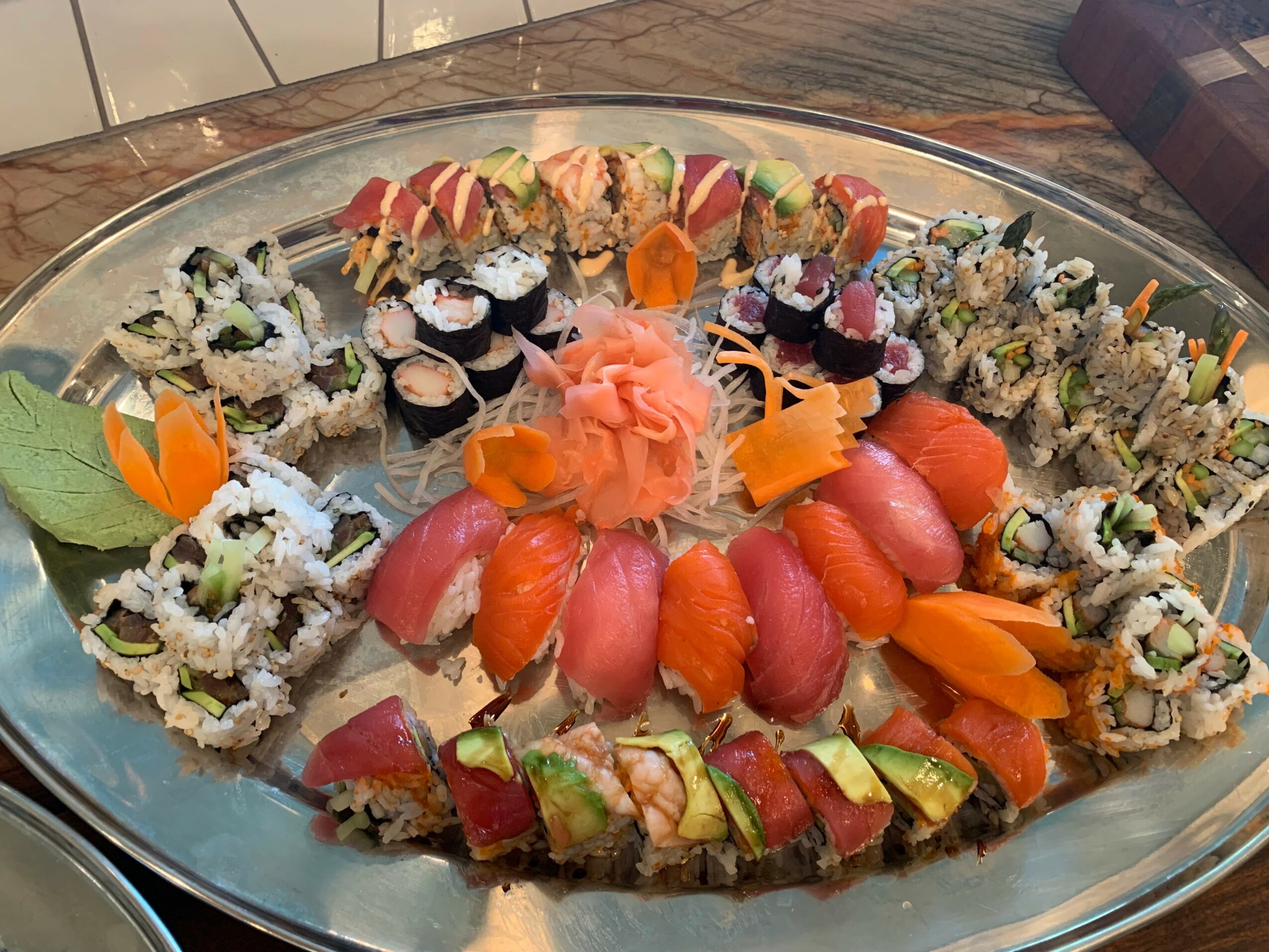Assorted sushi rolls and sashimi arranged on a silver platter with garnishes, including a pink flower and orange slices.