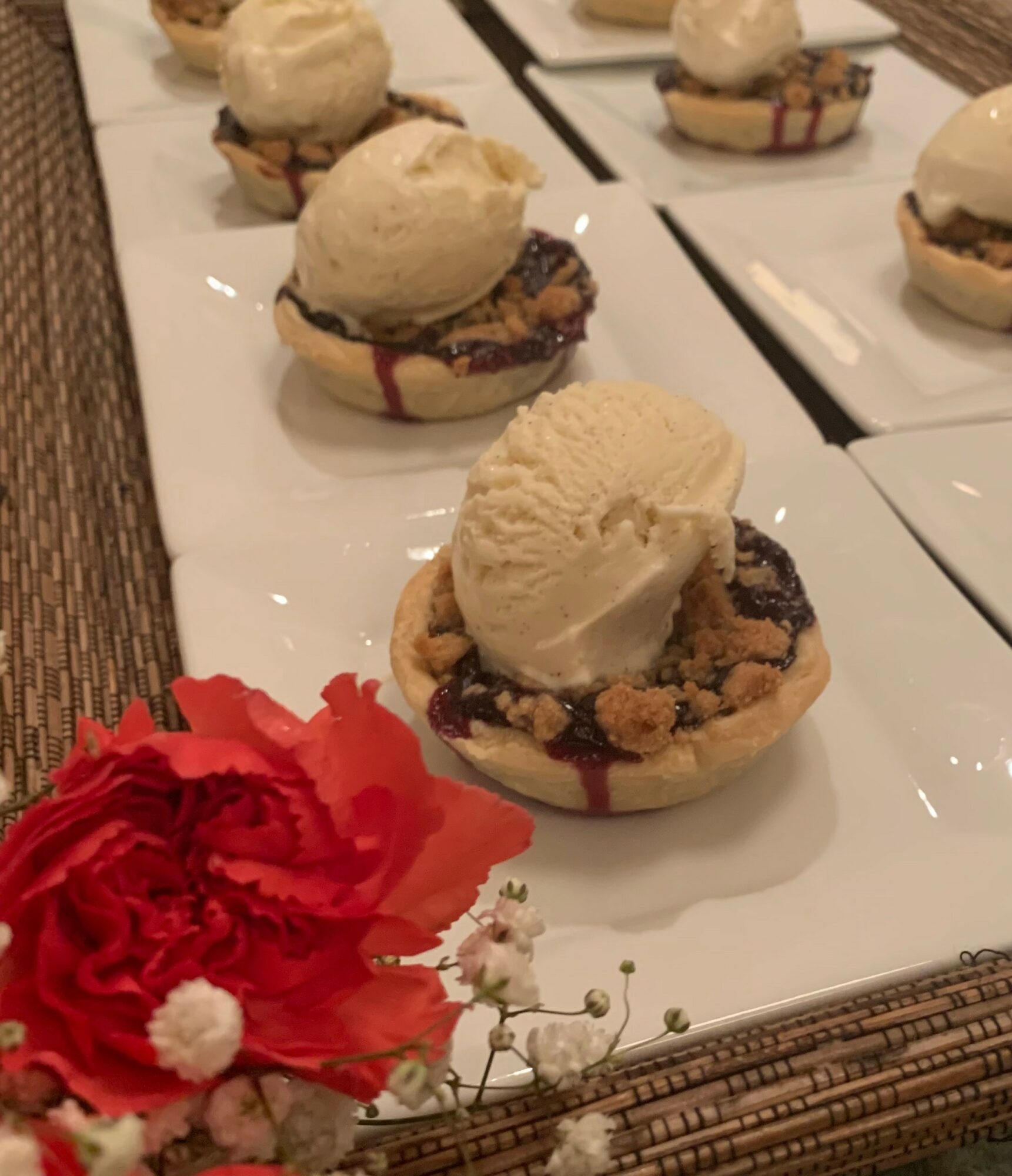 Multiple dessert cups with ice cream and toppings on white plates, with a red flower in the foreground.