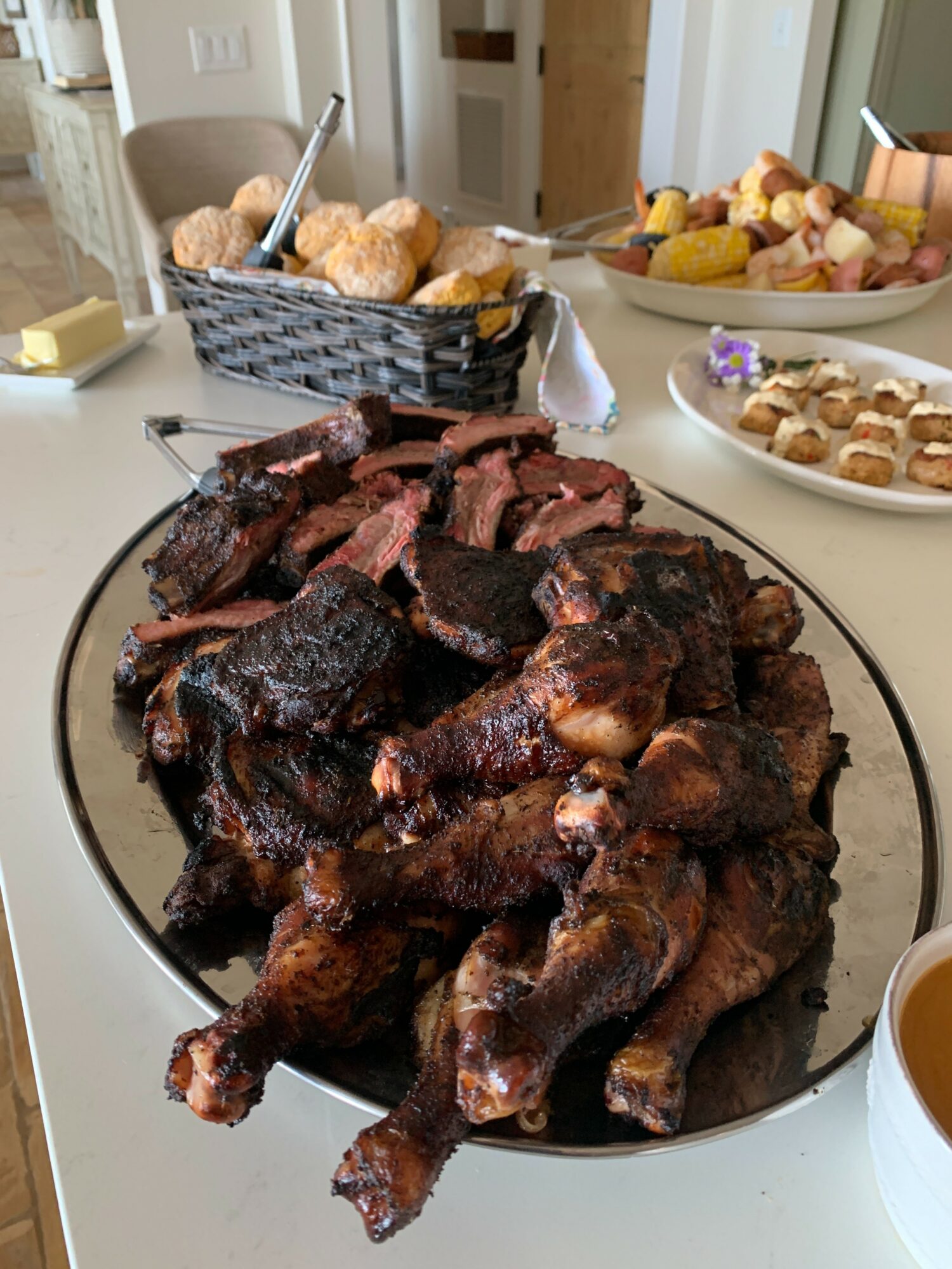 Plate of cooked chicken drumsticks and pieces on a white table with snacks and fruit in the background.