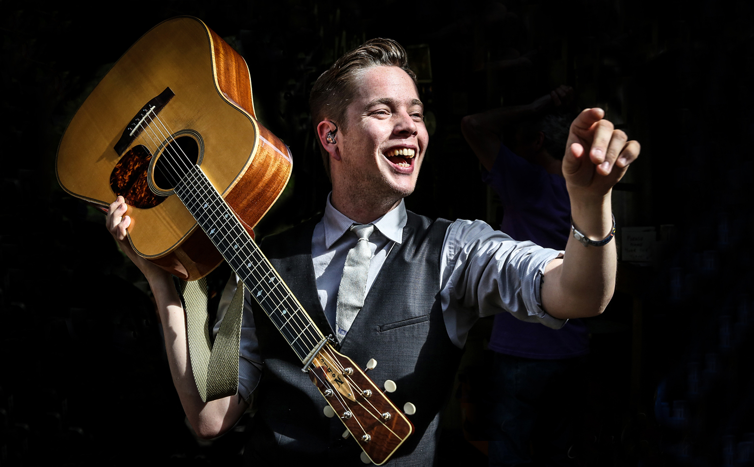 Person smiling, holding an acoustic guitar over shoulder, gesturing with hand, dark background