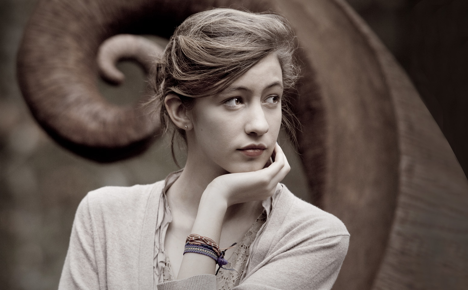A young woman with light hair resting her chin on her hand, looking to the side, with a large spiral-shaped object in the background.