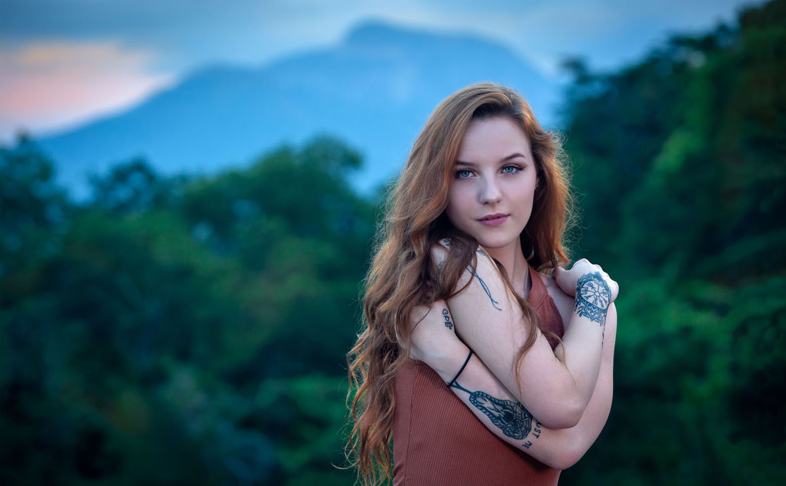 Young woman with long wavy hair standing outdoors with a blurred green forest background.