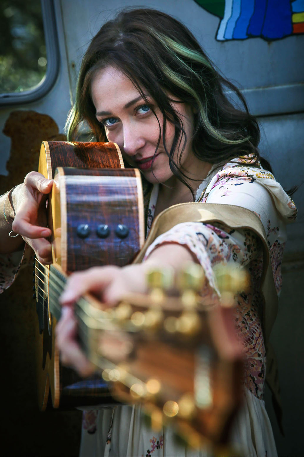 Young woman with wavy hair playing guitar, looking at camera, inside a vehicle with a window and colorful fabric, holding the guitar's neck.