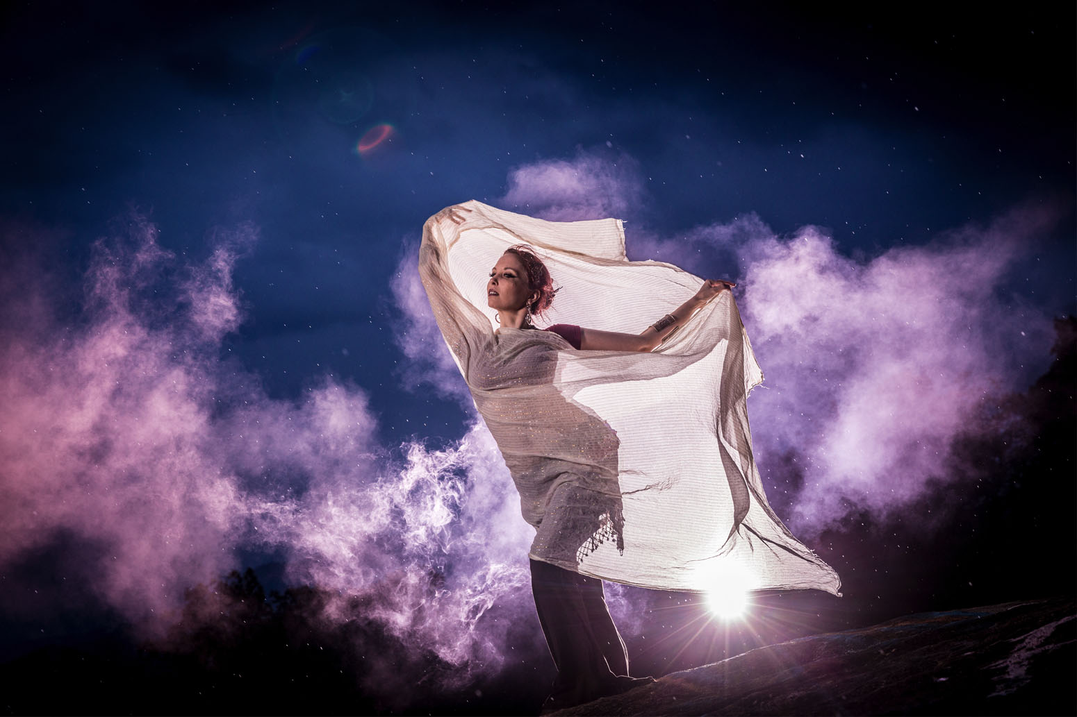 Pregnant woman holding a cloth against a starry sky with clouds and bright light behind her.
