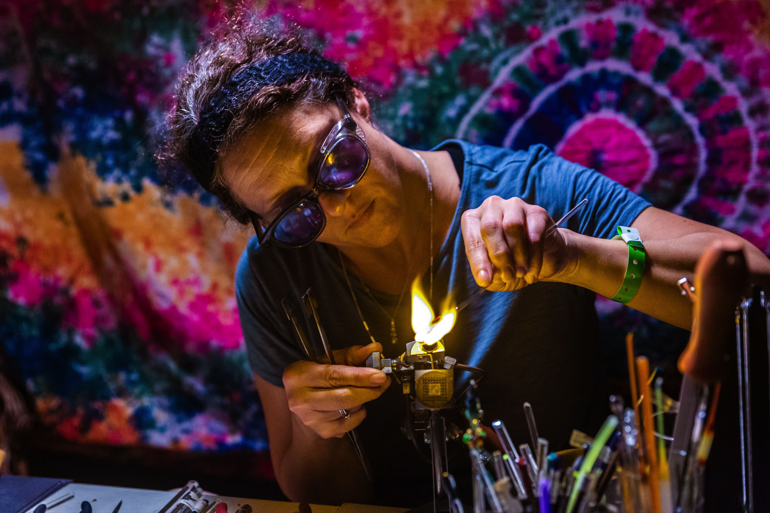 Person wearing sunglasses and a blue shirt, working with a flame and glass tools in a colorful studio, surrounded by glass rods.