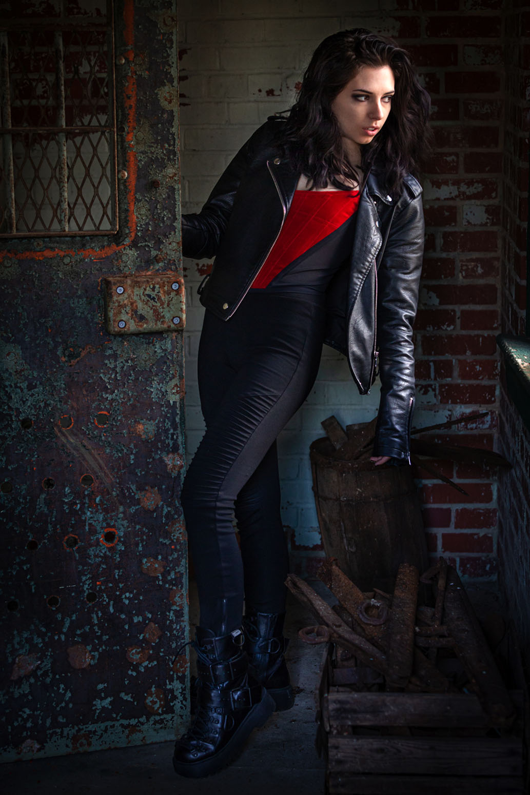 Young woman with dark curly hair in a leather jacket, red top, and jeans, leaning against a brick wall.
