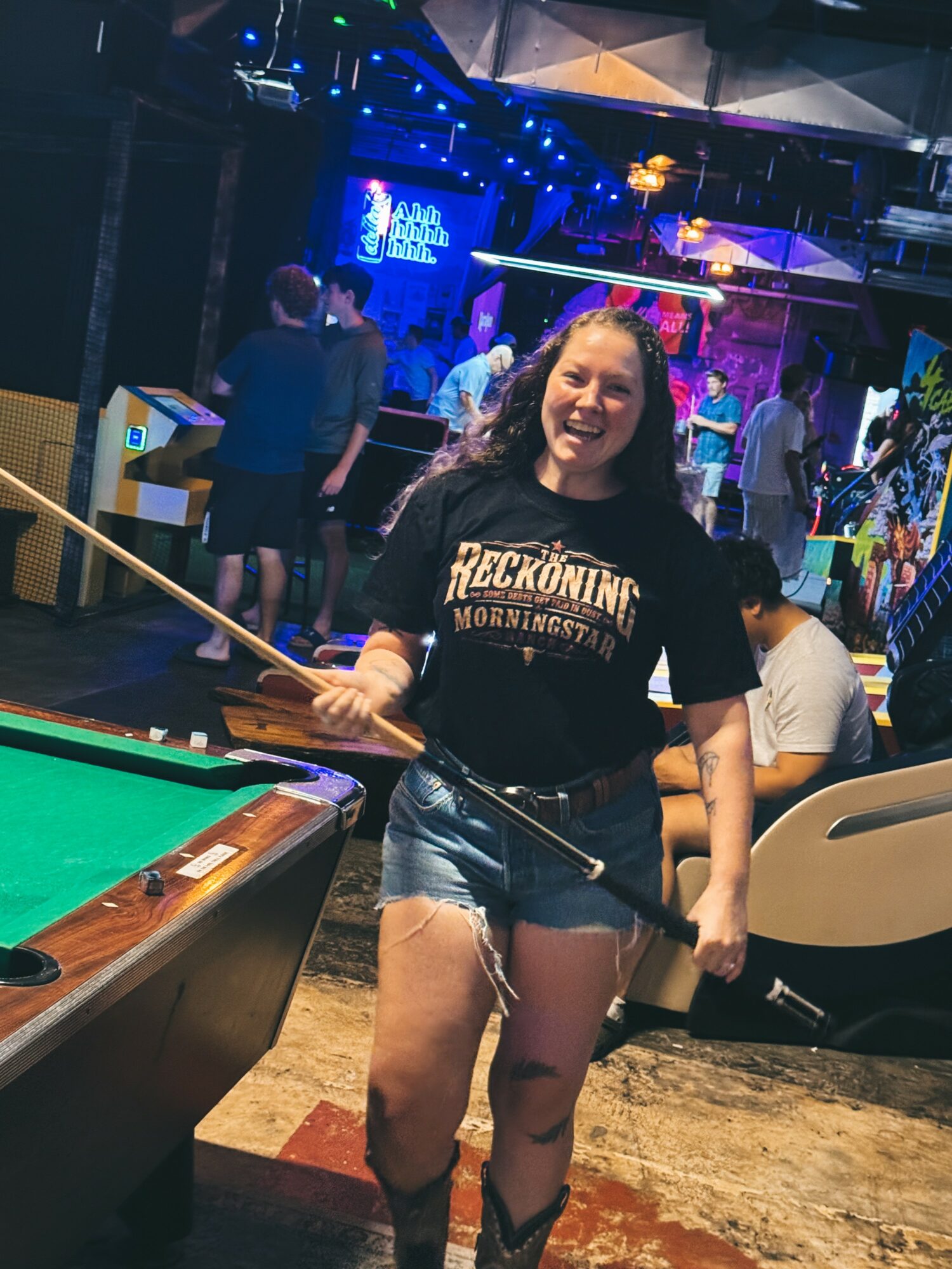 Young woman smiling, holding a pool cue, standing near a pool table in a lively bar with colorful lights and people in the background.