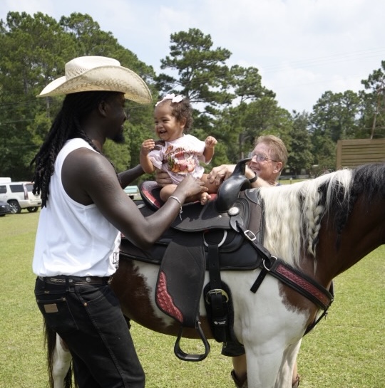 Woman in cowboy hat holding a smiling child on a horse, with a person beside the horse, outdoors with trees.