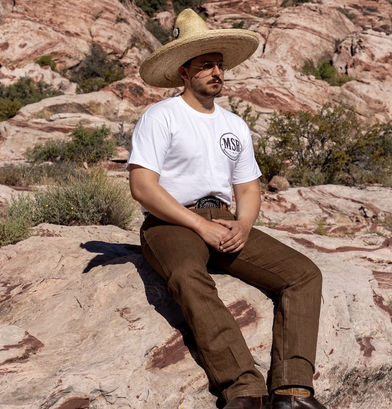 Man sitting on rocks in desert landscape, wearing a large straw hat, sunglasses, white t-shirt, and brown pants.