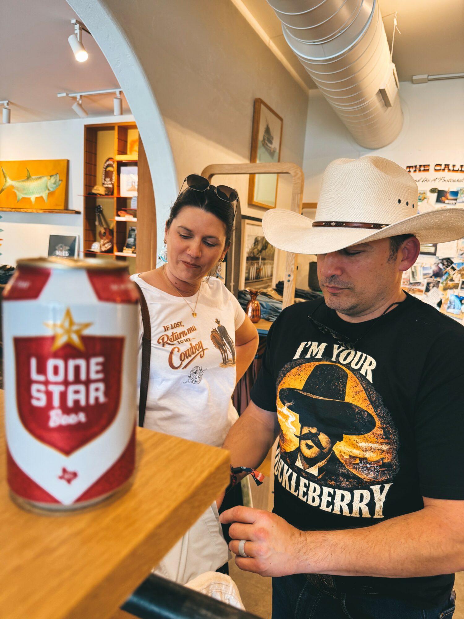 Two people in a store, one wearing a cowboy hat and a graphic T-shirt, the other in a white shirt, near a can of Lone Star beer.
