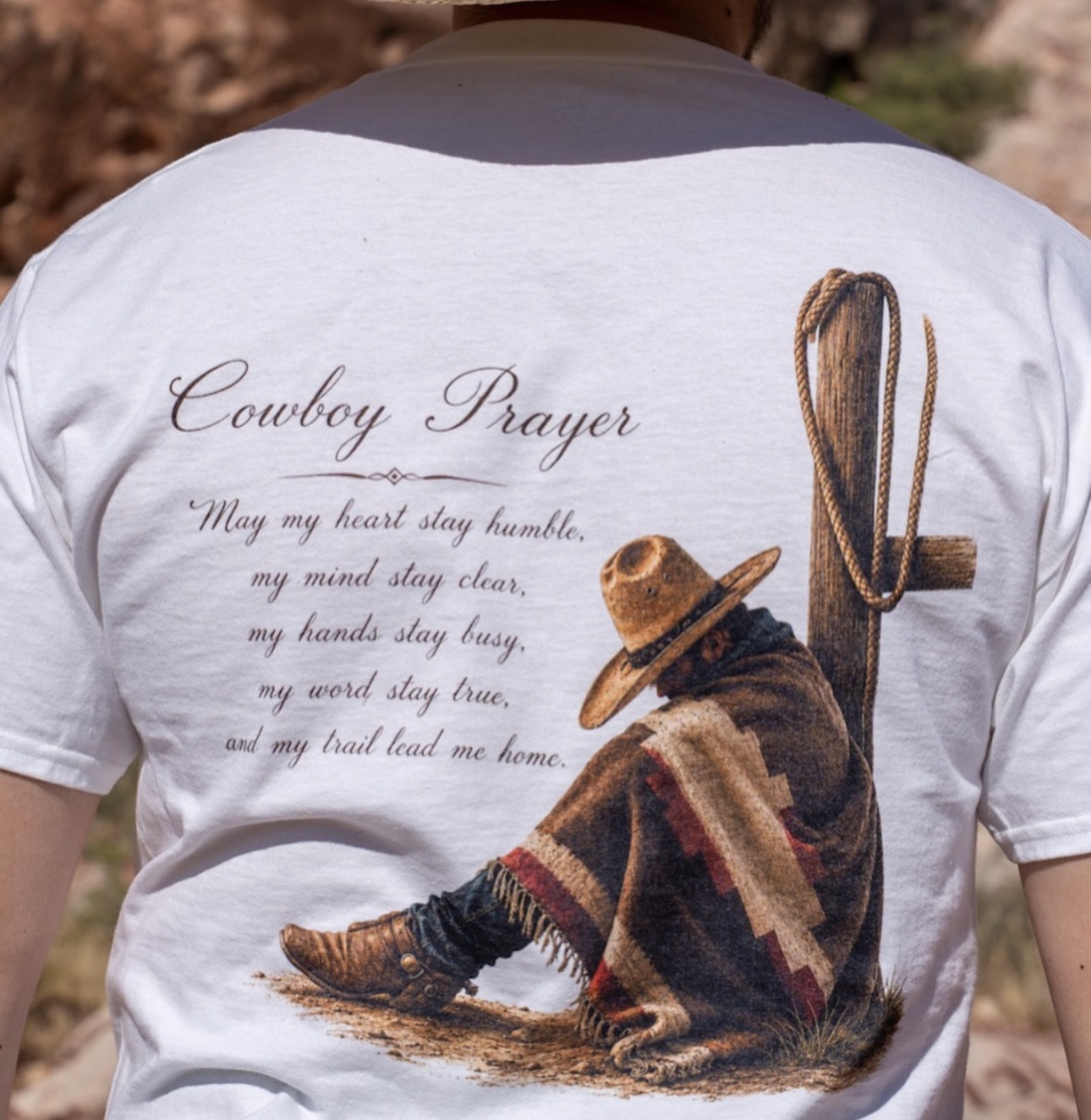 Person wearing a white T-shirt with a cowboy sitting on the ground, wearing a hat and blanket, with text about cowboy prayer.