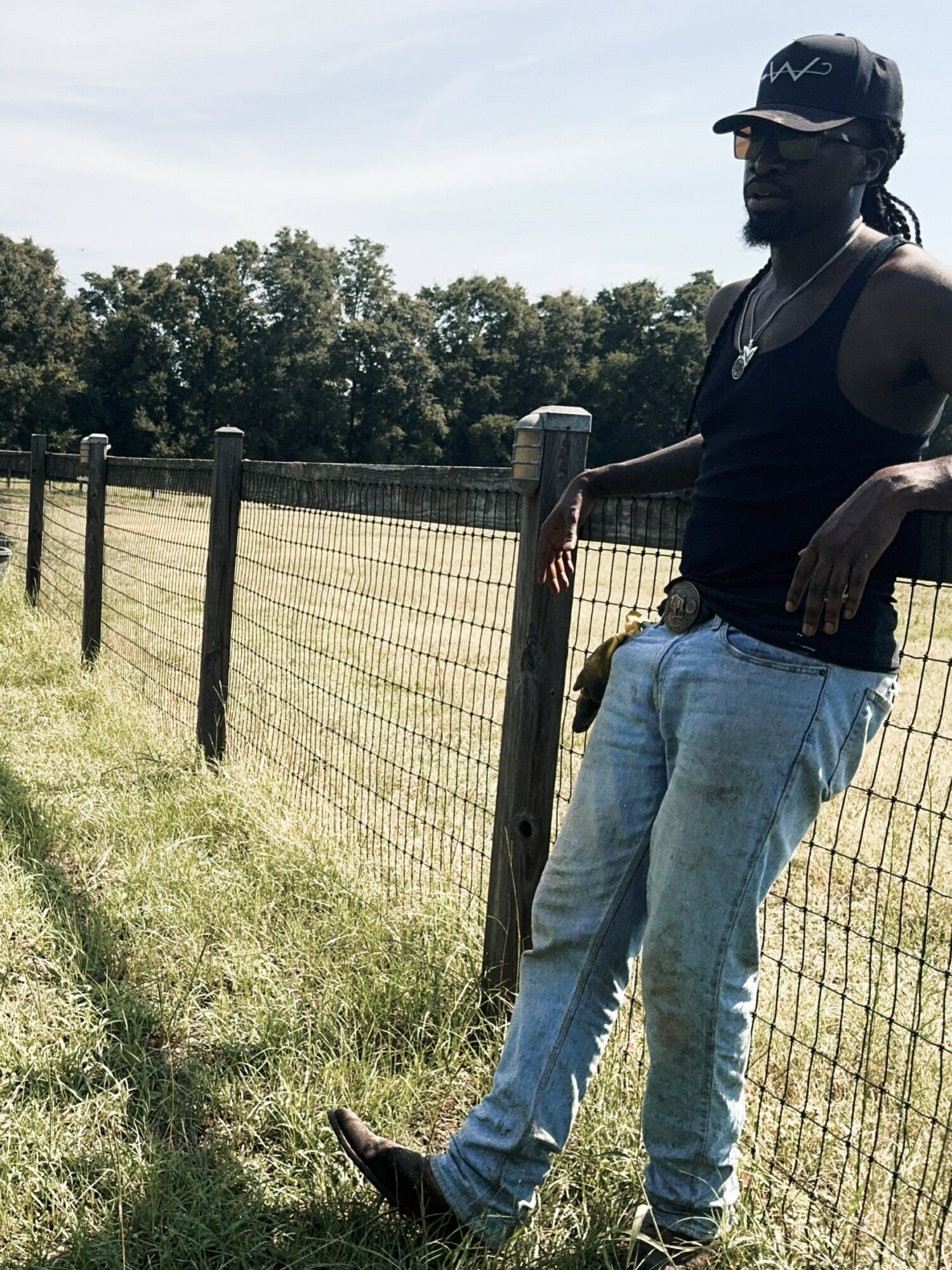 Person standing outdoors near a wire fence, wearing a cap, sunglasses, tank top, and jeans, with trees in the background.