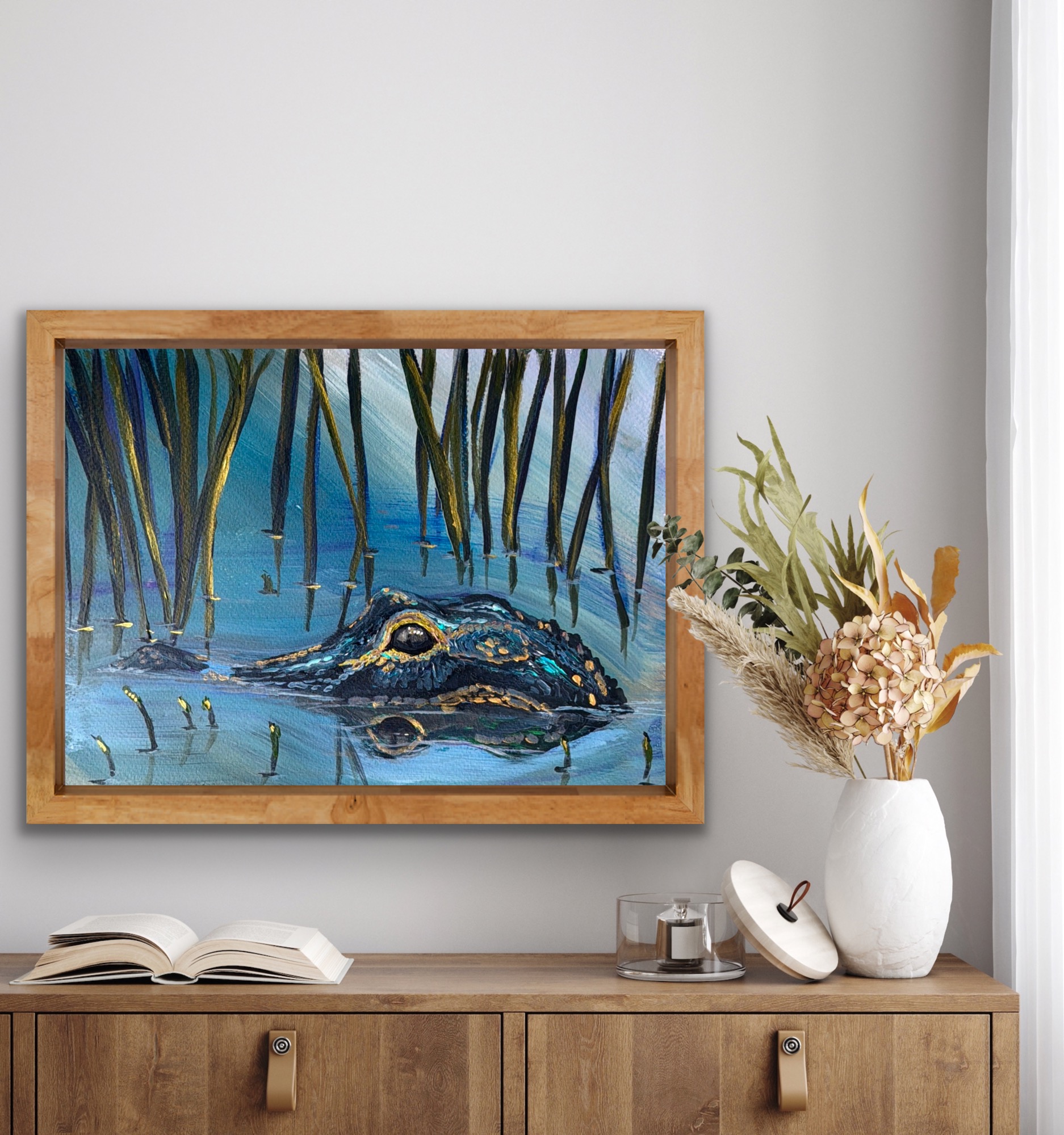 Painting of a crocodile in water with trees in the background, on a wooden shelf with a vase of dried flowers and an open book.