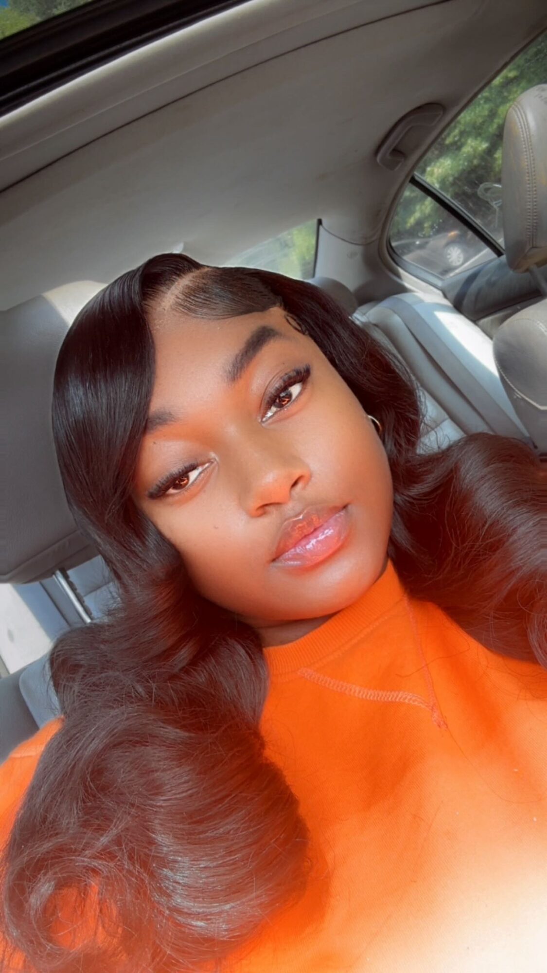 Young woman with long, wavy hair and makeup, wearing an orange top, sitting in a car, looking at the camera.