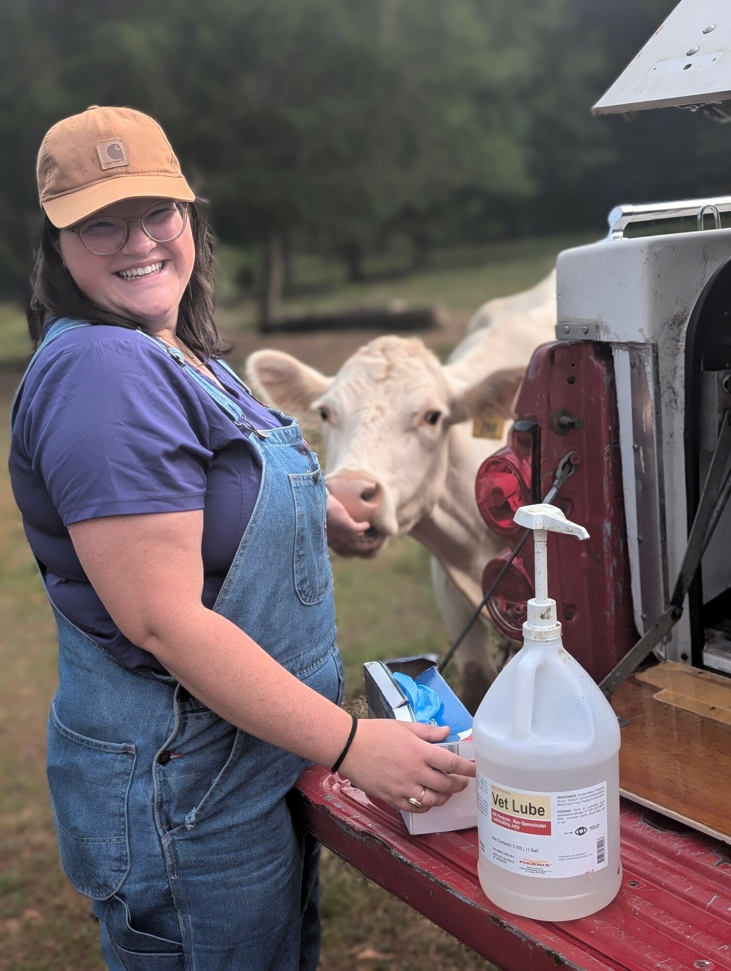 Smiling woman in blue overalls and a tan cap standing outdoors near a cow, with a spray bottle on a red table.