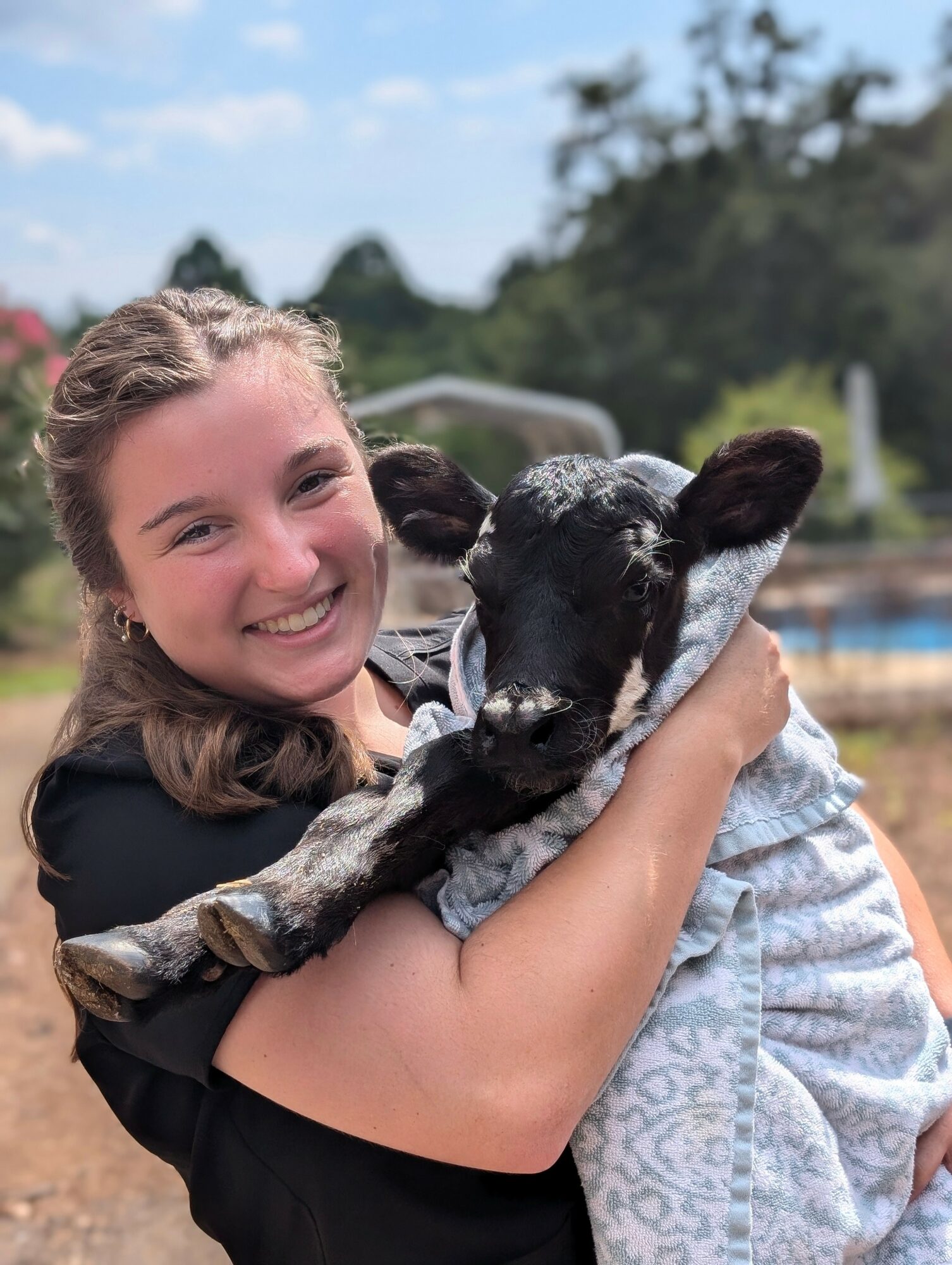 Girl smiling and hugging a black and white goat outdoors, trees and sky in background.