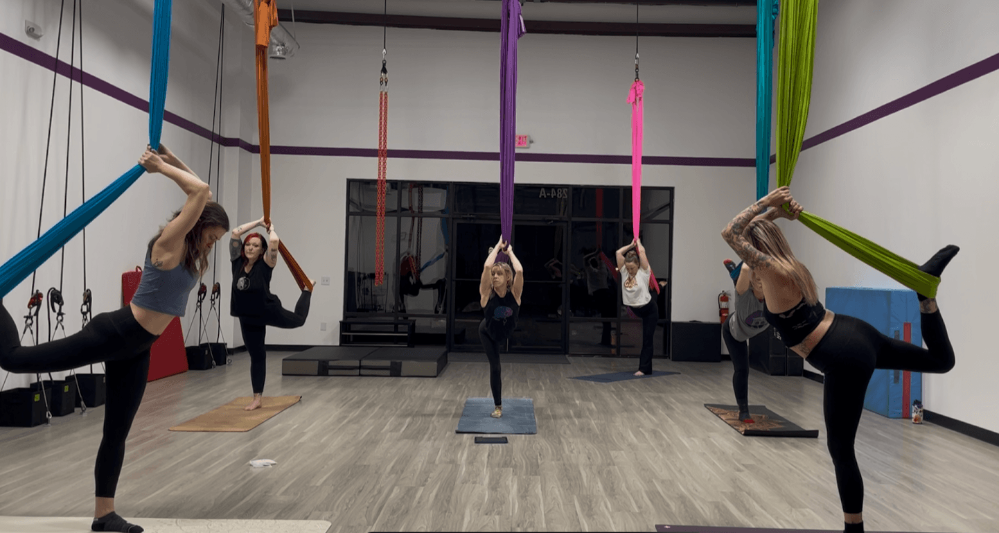 Group of people practicing aerial yoga with colorful hammocks in a studio, holding poses on mats.