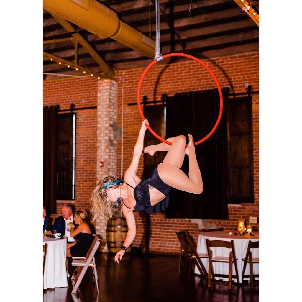 A woman performs aerial acrobatics on a red hoop hanging from the ceiling in a brick-walled venue.