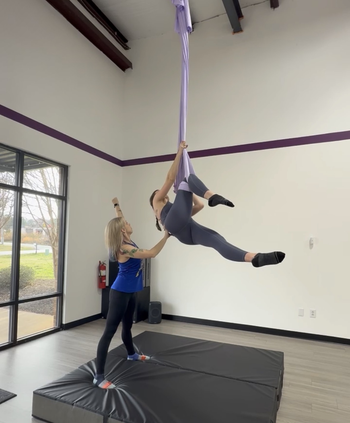 Person hanging from purple fabric in a gym, another person supporting them on a padded mat, large window in background.