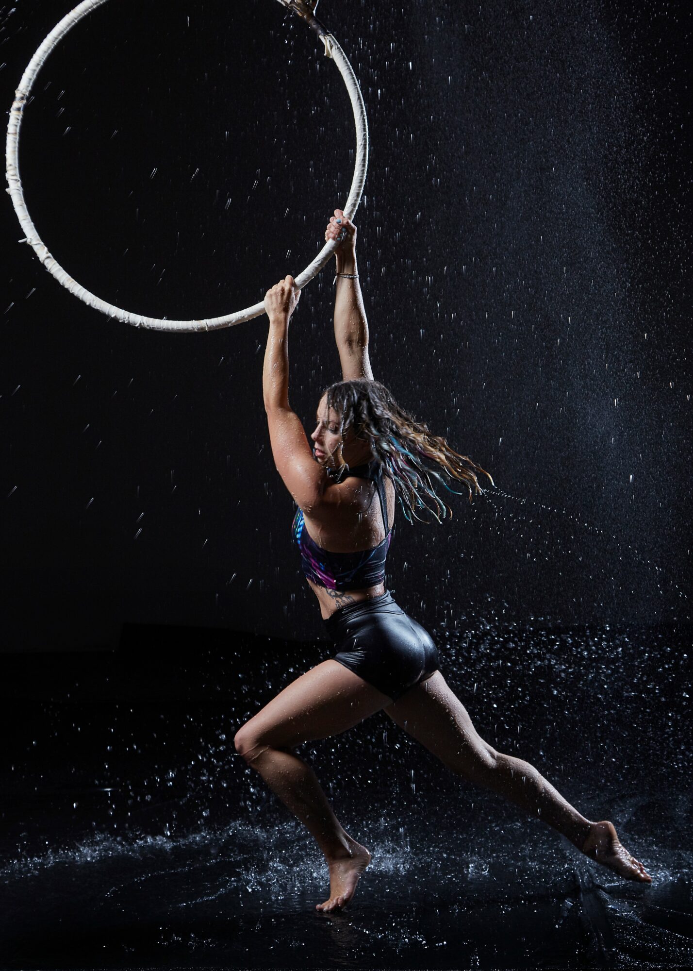 A woman holds a large hoop above her head, water splashes around her, and she is in a dynamic pose.