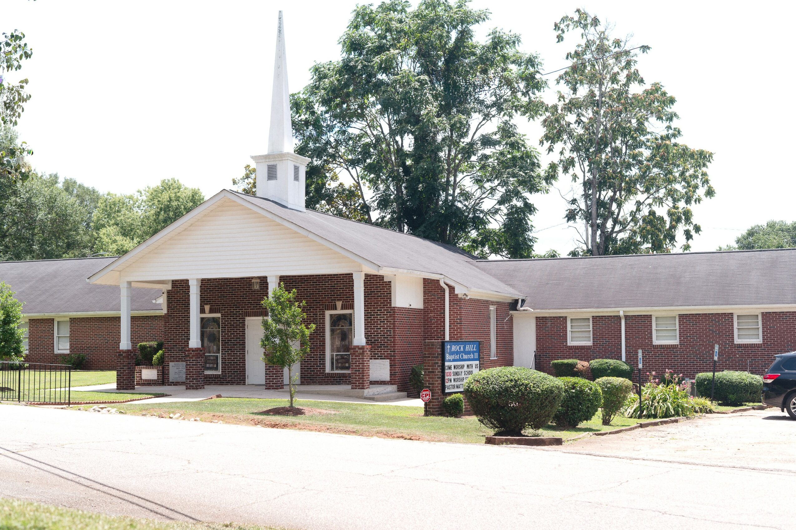 Brick church with white steeple, surrounded by trees and bushes, parking lot in front, clear sky.