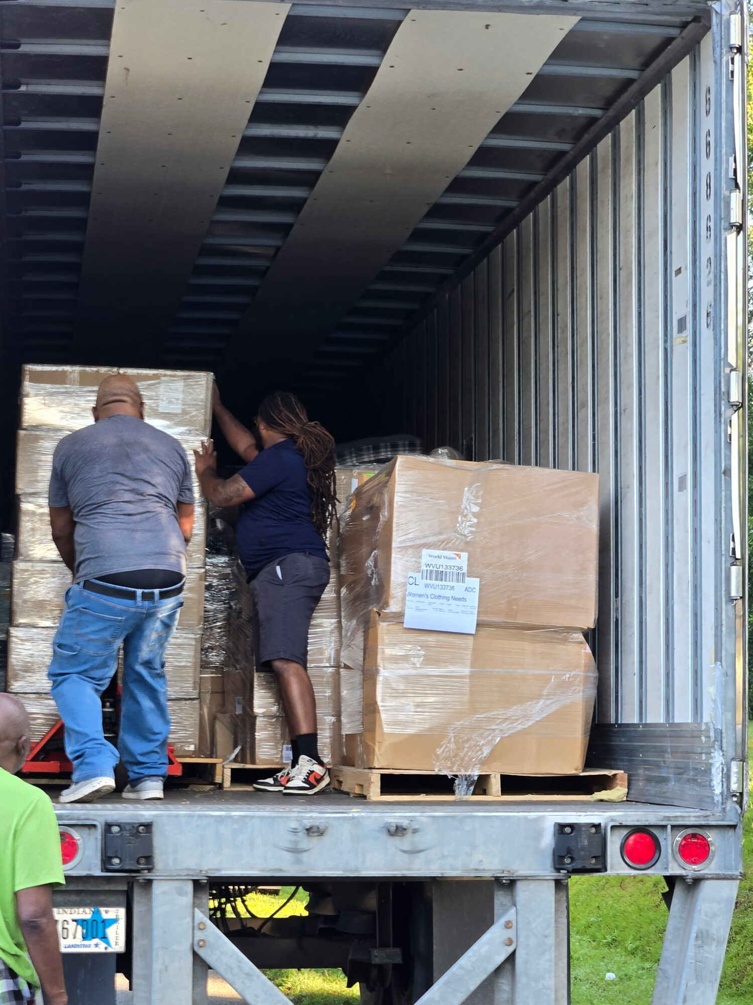 Two people stand inside a truck with boxes on pallets, loading or unloading items, with a person in a green shirt partially visible in the foreground.